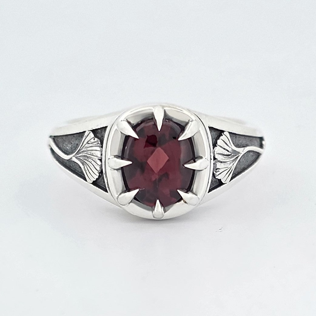 Sterling silver ring featuring an oval deep-red garnet in a claw setting, with sculpted ginkgo leaf details on oxidised shoulders.