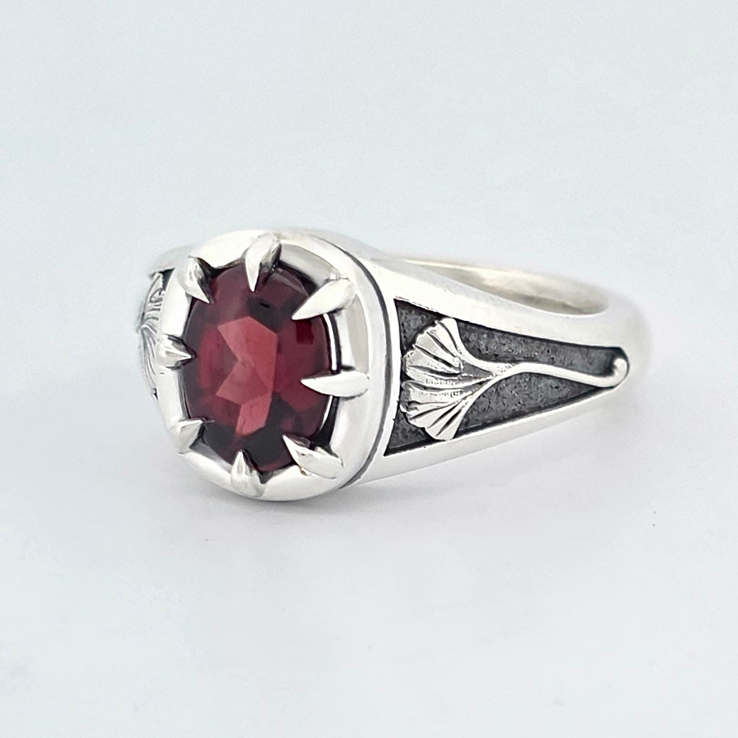 Sterling silver ring featuring an oval deep-red garnet in a claw setting, with sculpted ginkgo leaf details on oxidised shoulders.