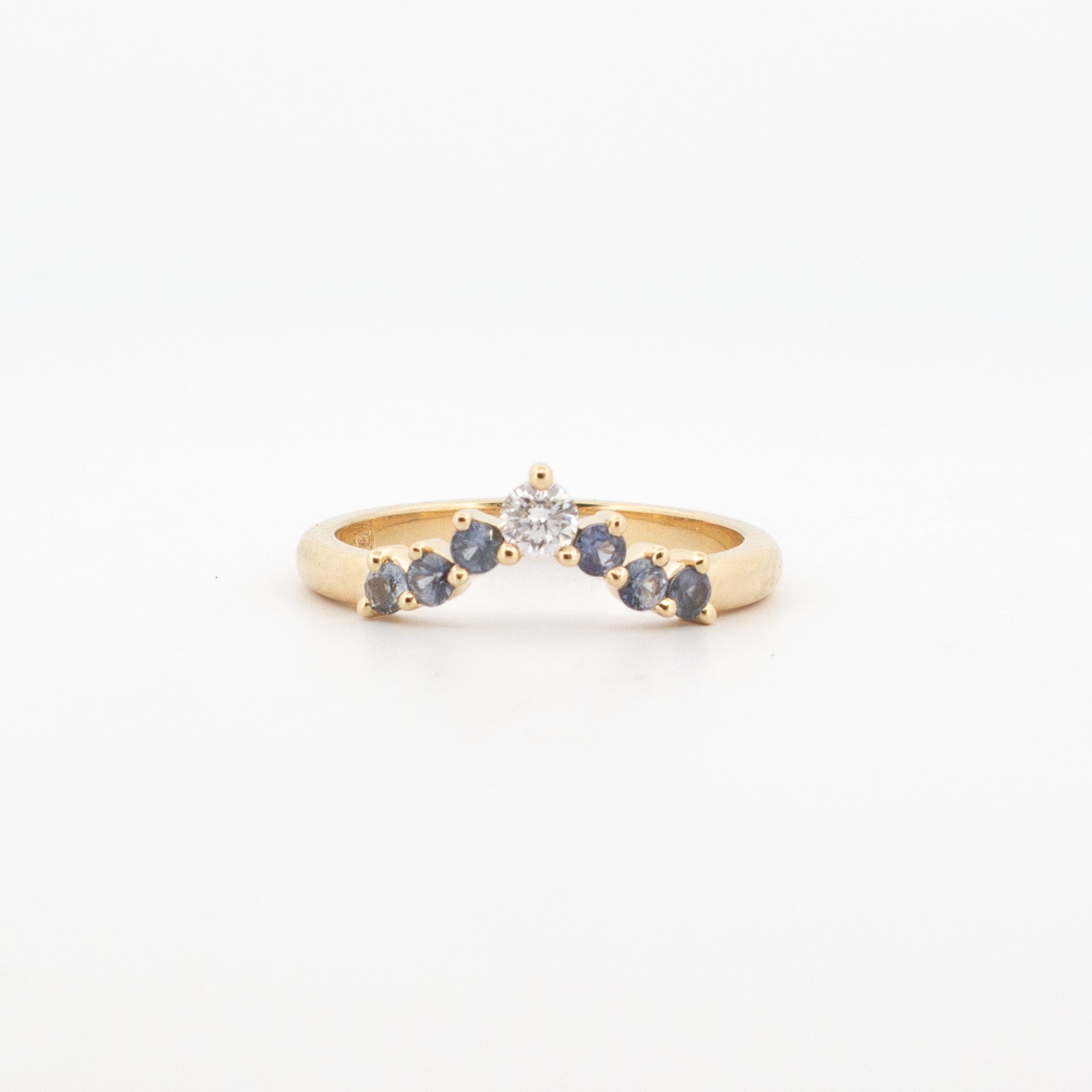 Front view of an 18ct yellow gold chevron-shaped ring featuring a central round white diamond flanked by a symmetrical gradient of prong-set blue and grey diamonds. The stones taper in size toward the shoulders, creating a soft fading effect along the V-shaped band. The ring has a polished finish and a low-profile setting, designed for stacking or as a contoured wedding band with a modern, geometric form.