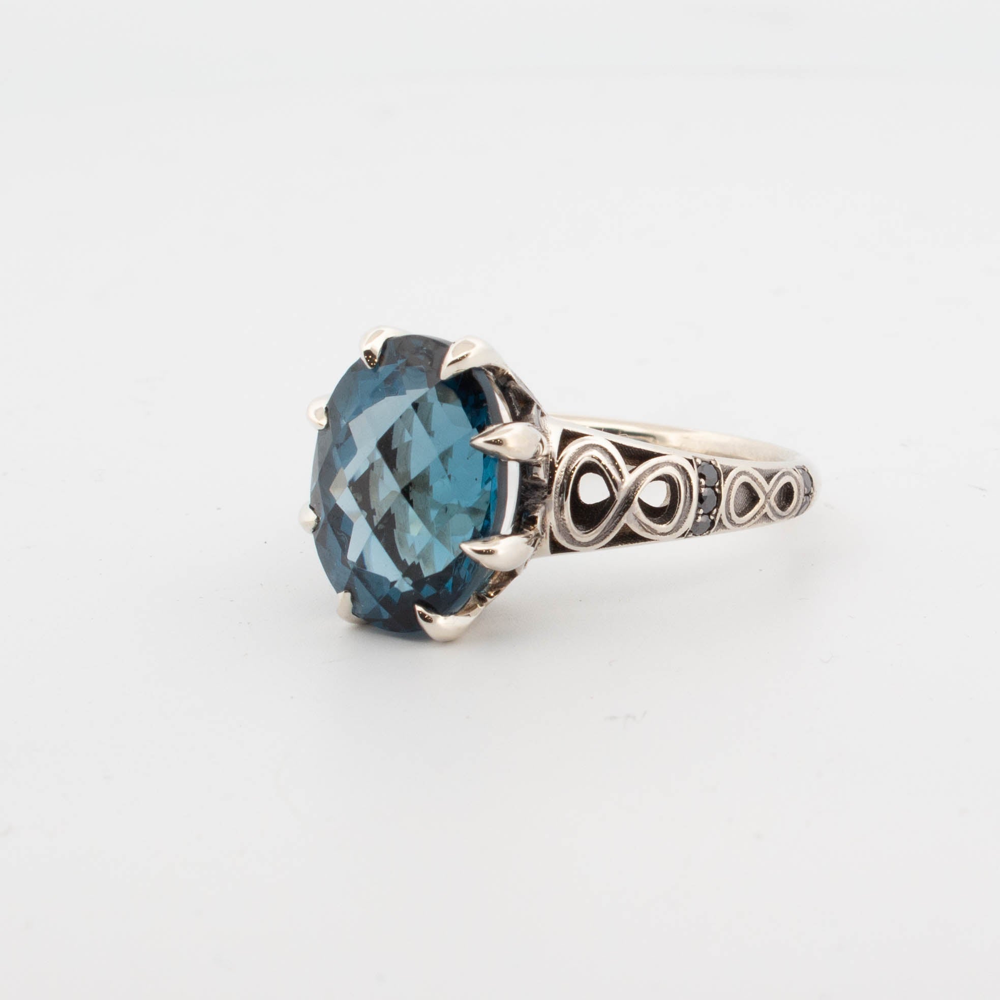 9ct white gold ring featuring a large oval-cut London Blue Topaz in a claw setting, with infinity symbol openwork shoulders and pavé-set black diamonds on the band.