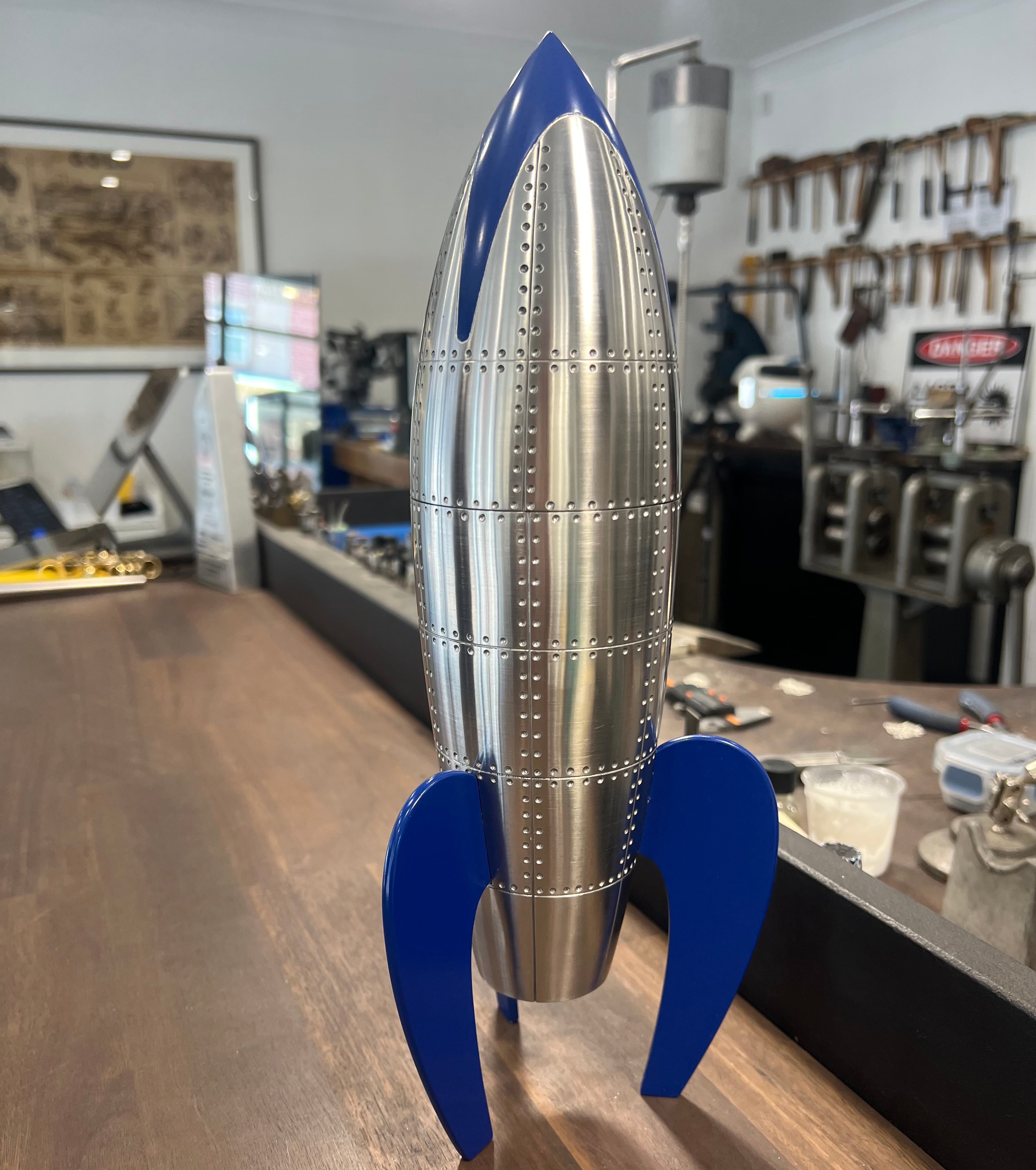 Custom cremation urn designed in the form of a retro rocket, crafted from stainless steel and brass with rivet detailing, and finished with glossy blue enamel fins and nose cone; stands 34cm tall.