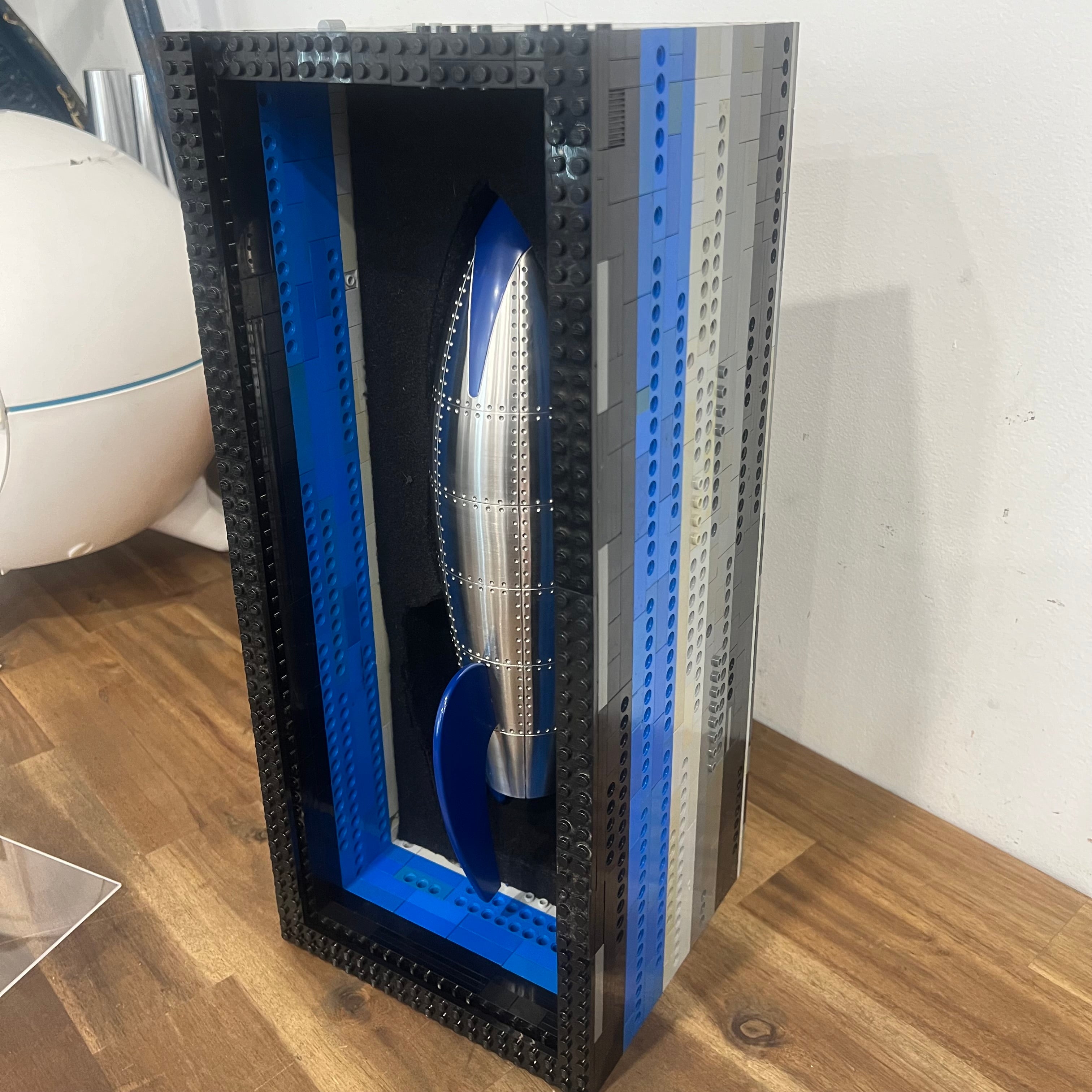 Custom cremation urn designed in the form of a retro rocket, crafted from stainless steel and brass with rivet detailing, and finished with glossy blue enamel fins and nose cone; stands 34cm tall. encased in lego