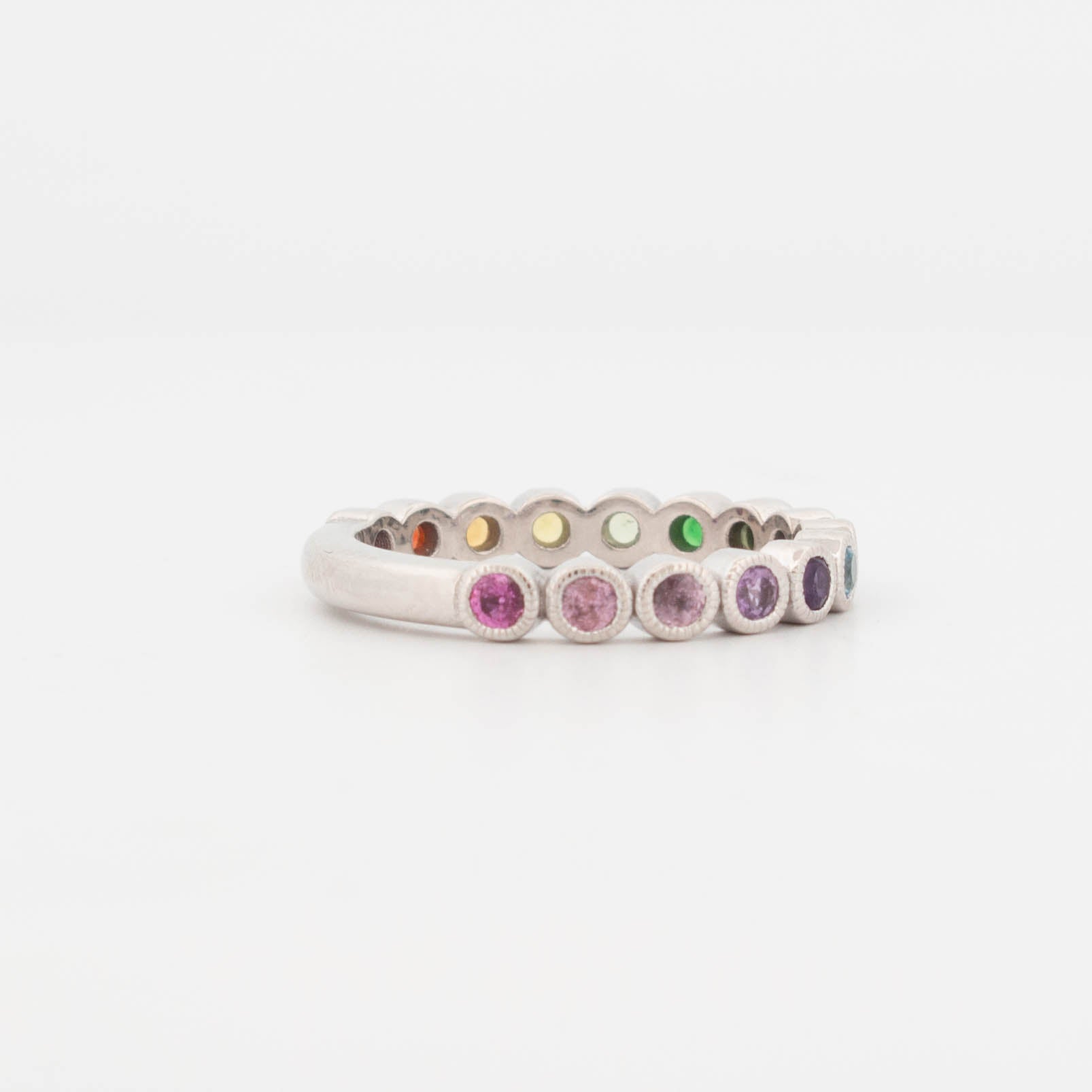 9ct white gold rainbow sapphire ring featuring a full circle of bezel-set round sapphires in a smooth colour-fade gradient—from deep green and lime to yellow, orange, red, and pink. Each gemstone is framed in a fine milgrain bezel, giving the ring a vintage-inspired finish with a modern twist. This eternity-style band showcases a vibrant spectrum of sapphires, ideal for stacking, pride jewellery, or as a colourful wedding band alternative.