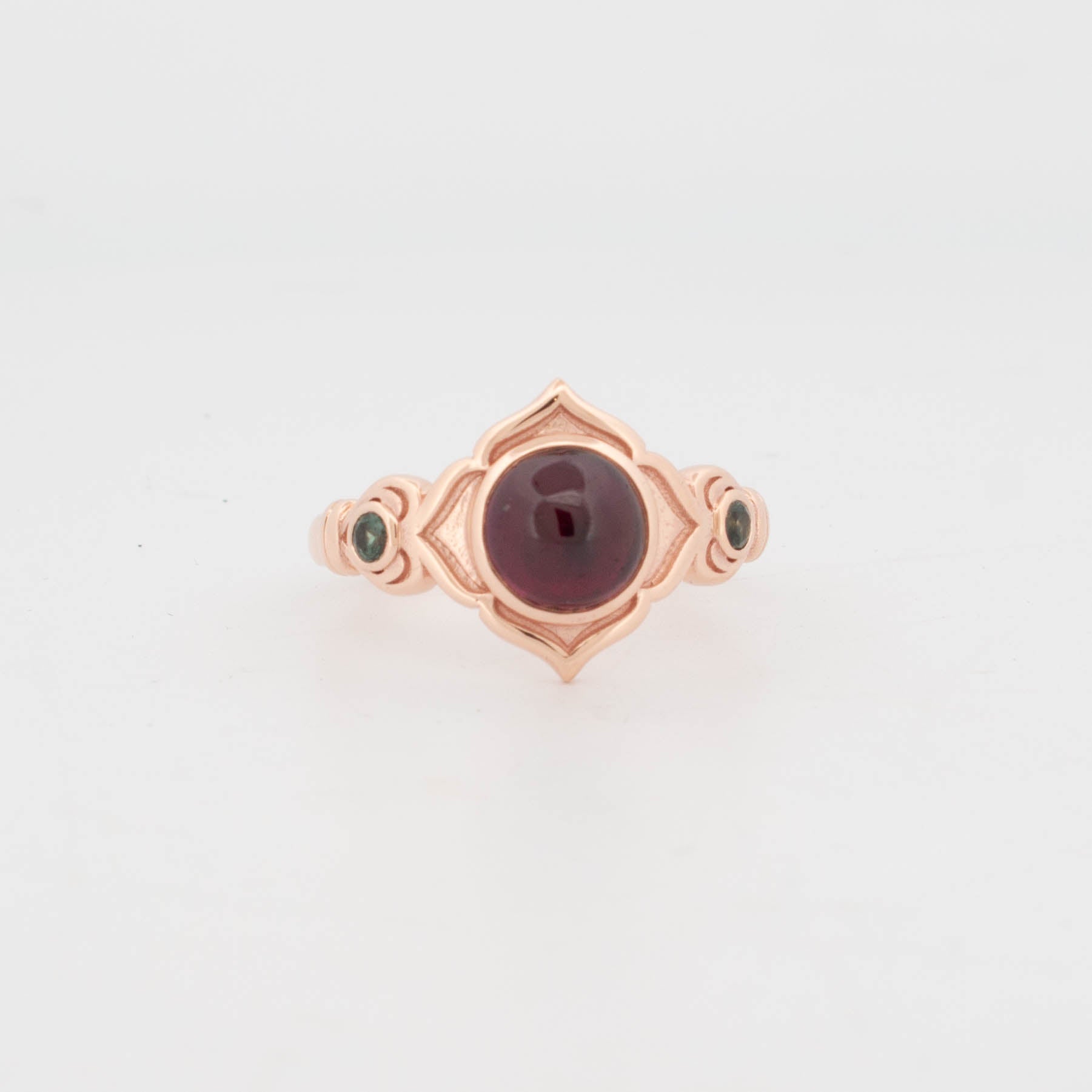 Front view of a 9ct rose gold ring featuring a central round cabochon-cut rhodolite garnet in a bezel setting, framed by a symmetrical pointed mandala-style motif. The shoulders are accented with flush-set round tsavorite garnets, adding a contrasting green hue. The ring has a polished finish with intricate architectural detailing, combining Art Deco and antique-inspired design elements.