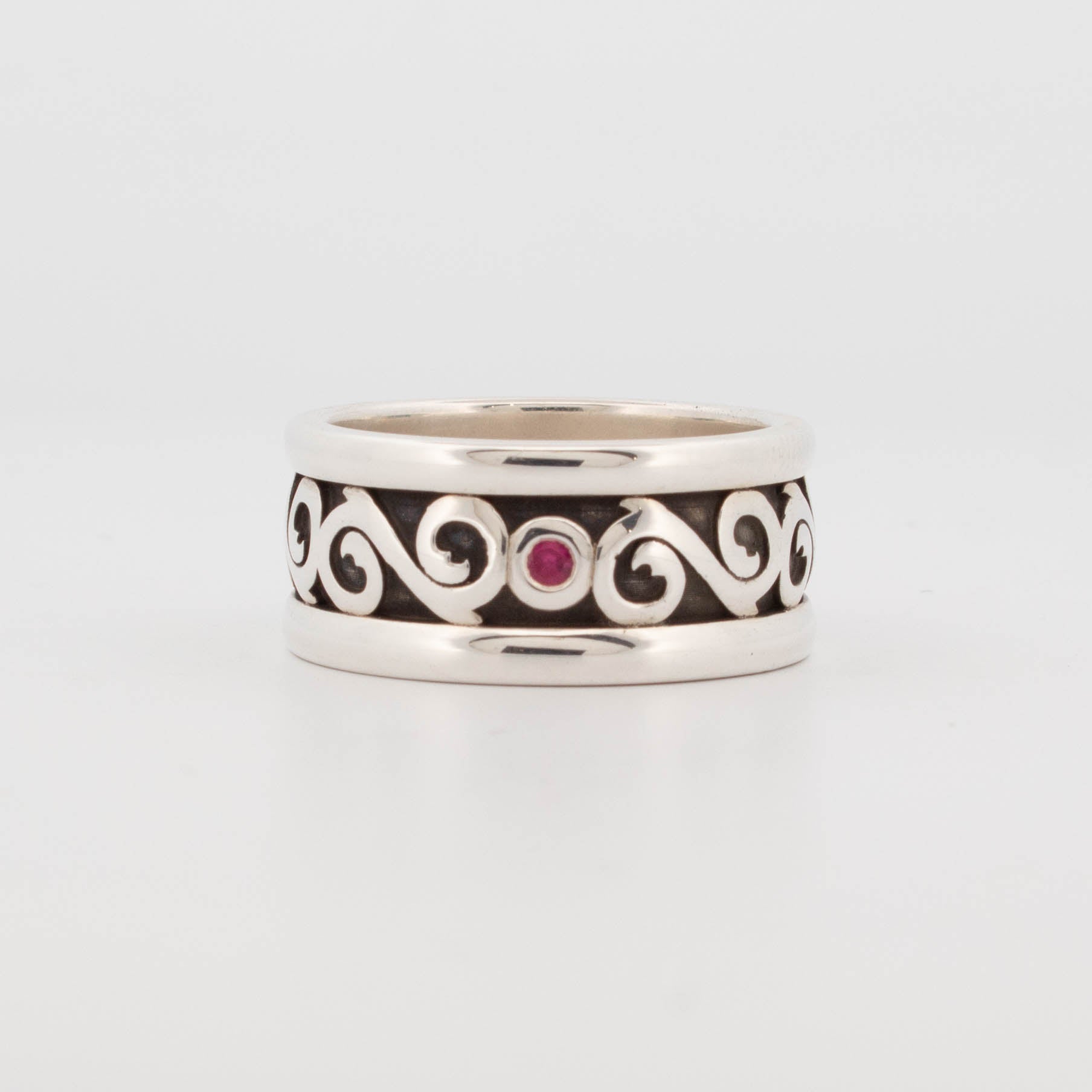 10mm wide sterling silver band ring featuring raised scrolling filigree on an oxidised background, set with a central round ruby; handcrafted gothic-style ring with baroque detailing.