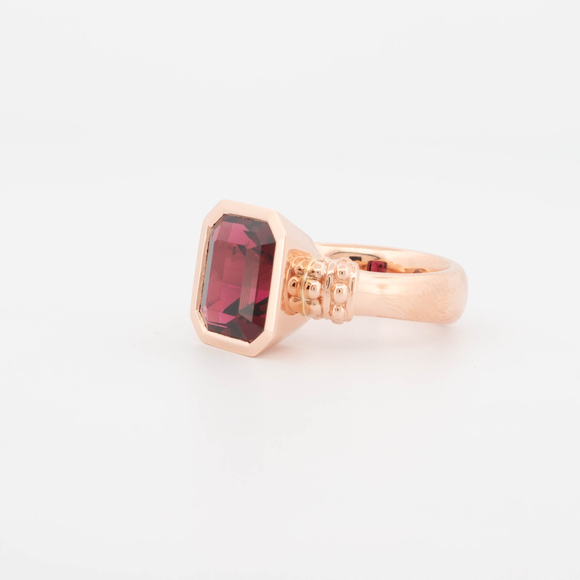 18ct rose gold ring featuring a richly coloured emerald-cut Rhodolite Garnet in a bezel-style setting, with a bold, polished band and decorative beaded shoulder detail.