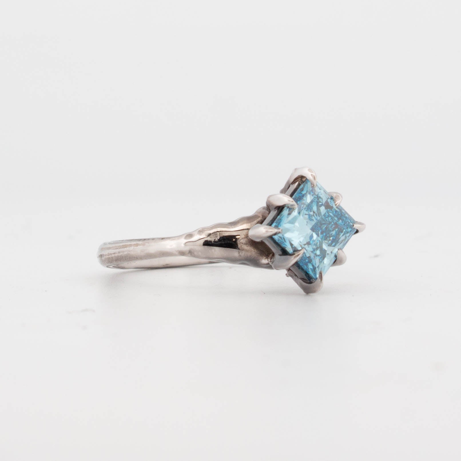Side view of 9ct white gold ring featuring a princess-cut blue diamond held in claw-like talon prongs, set on an organically textured band; handcrafted fantasy-inspired design with a bold gemstone centre.