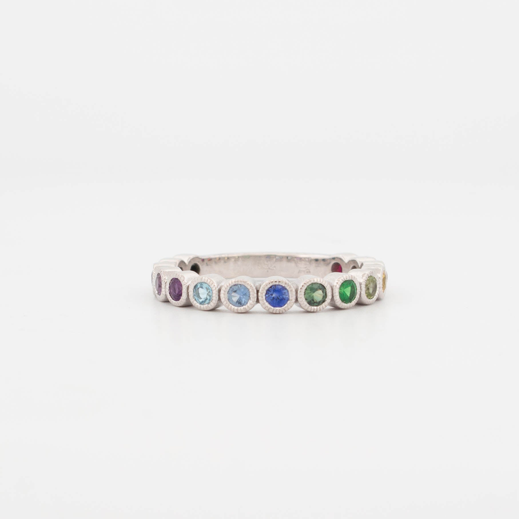 9ct white gold rainbow sapphire ring featuring a full circle of bezel-set round sapphires in a smooth colour-fade gradient—from deep green and lime to yellow, orange, red, and pink. Each gemstone is framed in a fine milgrain bezel, giving the ring a vintage-inspired finish with a modern twist. This eternity-style band showcases a vibrant spectrum of sapphires, ideal for stacking, pride jewellery, or as a colourful wedding band alternative.