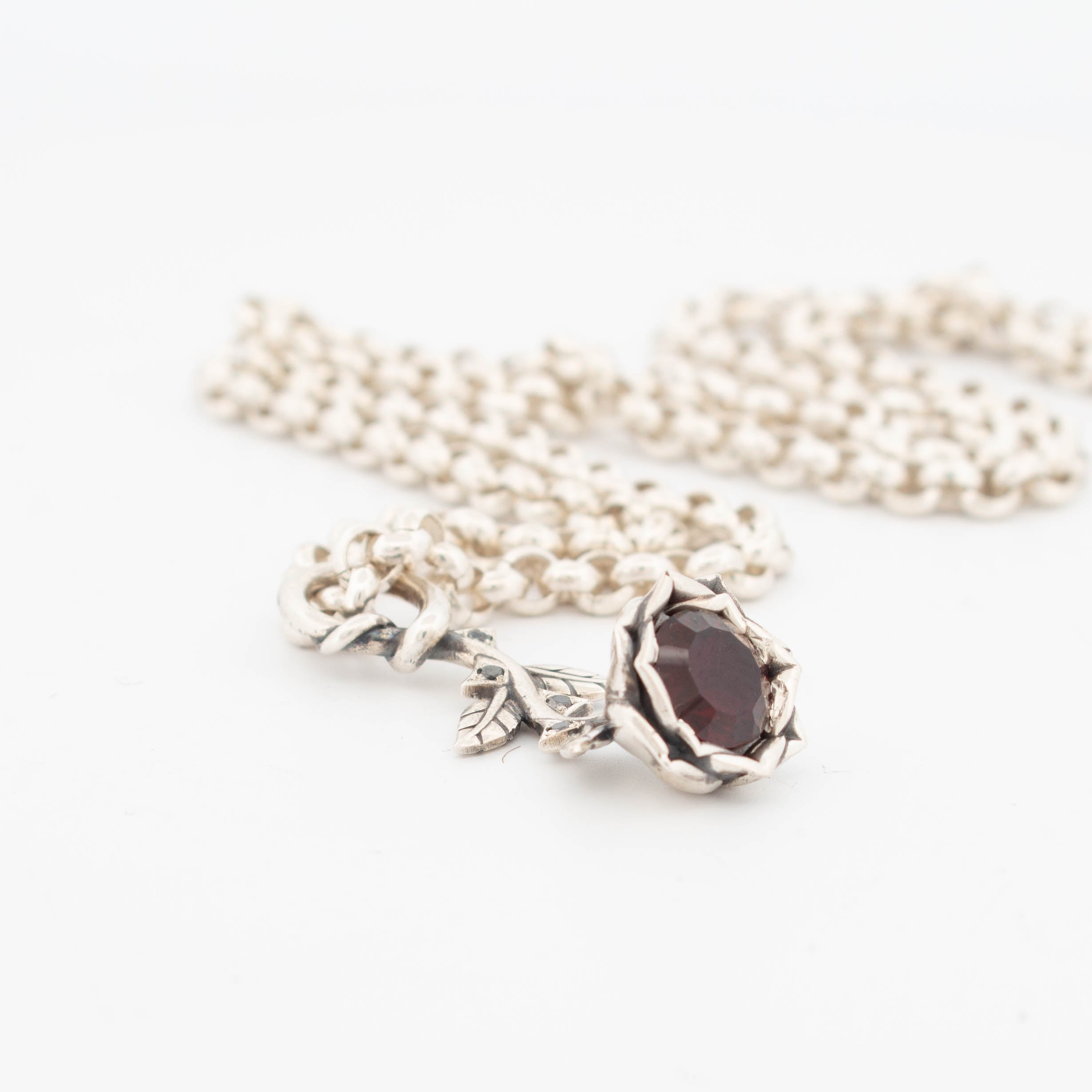 Sterling silver rose pendant necklace featuring a deep red garnet at the centre of a sculpted bloom, accented with black diamonds in the leaves, suspended from a chunky silver chain; handcrafted gothic floral jewellery with gemstone detailing.