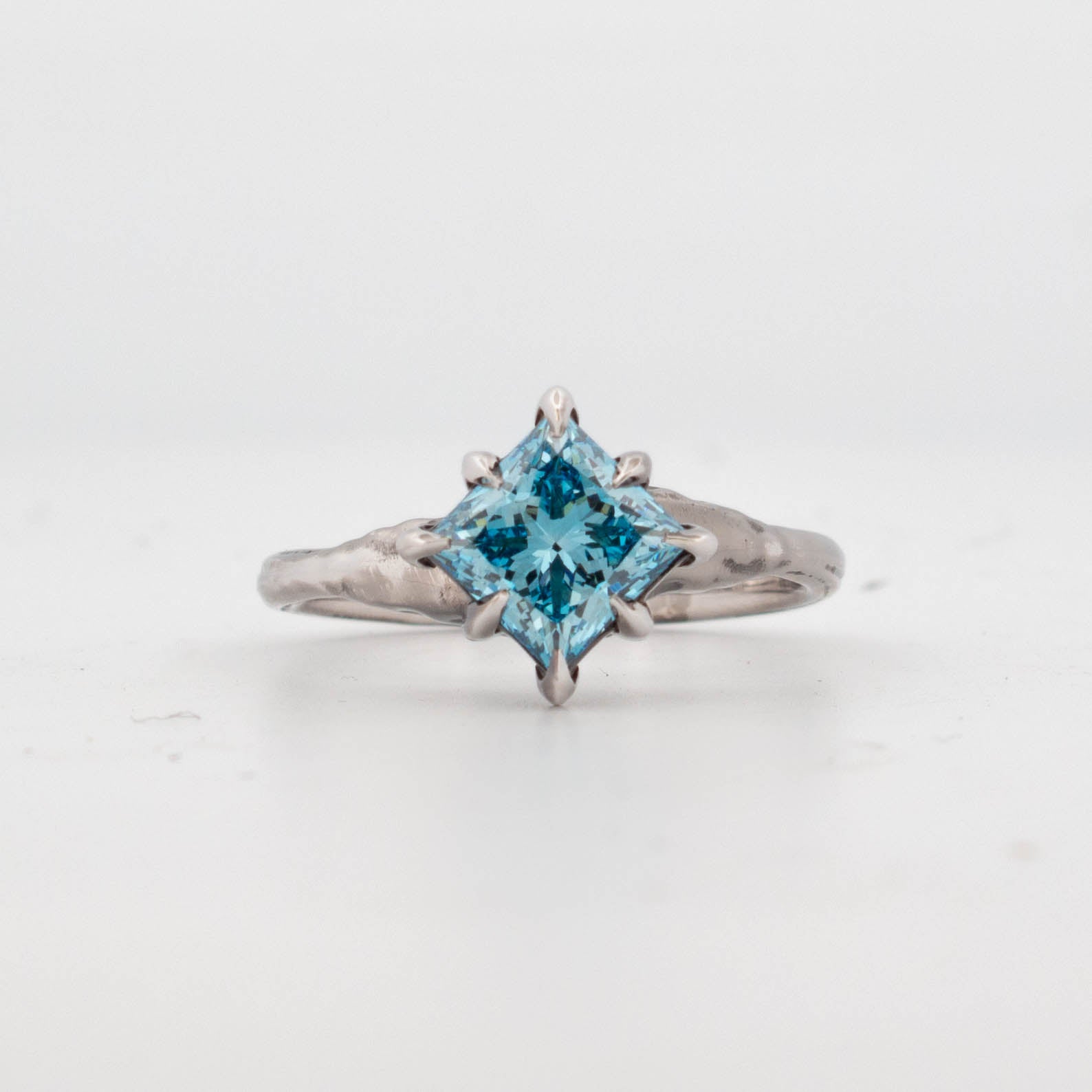 Front view of 9ct white gold ring featuring a princess-cut blue diamond held in claw-like talon prongs, set on an organically textured band; handcrafted fantasy-inspired design with a bold gemstone centre.