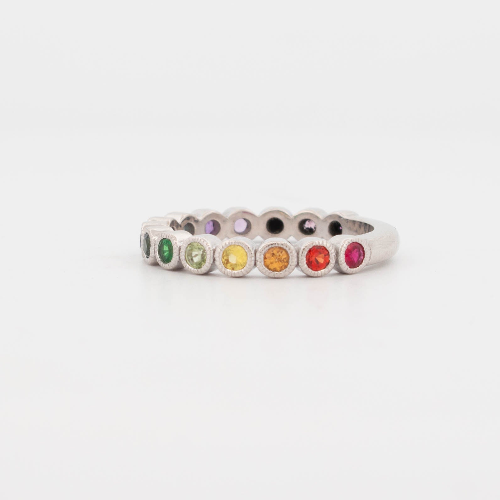 9ct white gold rainbow sapphire ring featuring a full circle of bezel-set round sapphires in a smooth colour-fade gradient—from deep green and lime to yellow, orange, red, and pink. Each gemstone is framed in a fine milgrain bezel, giving the ring a vintage-inspired finish with a modern twist. This eternity-style band showcases a vibrant spectrum of sapphires, ideal for stacking, pride jewellery, or as a colourful wedding band alternative.