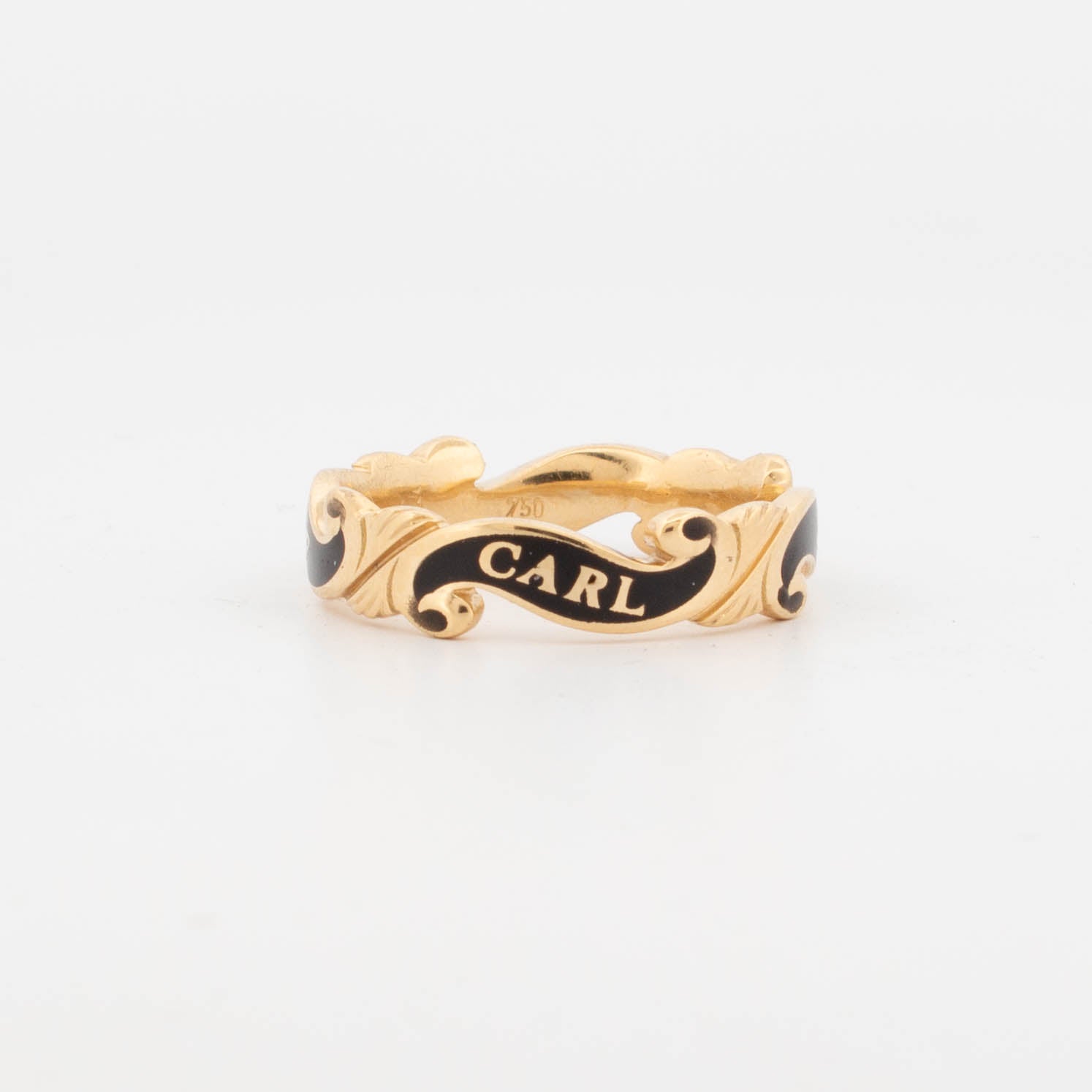 18ct yellow gold memorial ring with black enamel scrollwork, engraved with the inscription 'IN MEMORY OF CARL' around the band; designed as a tribute piece with ornate ribbon-like detailing.