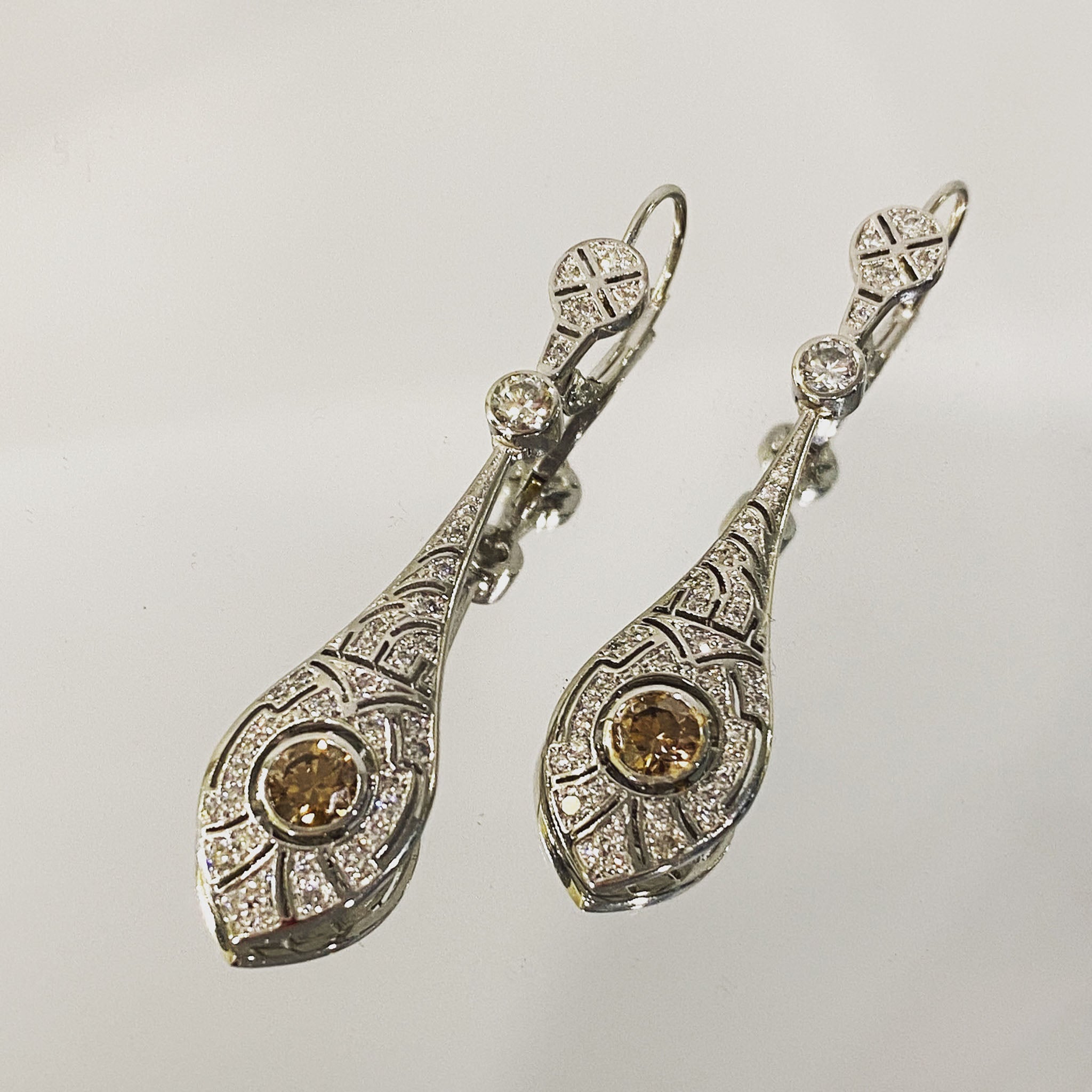 Platinum Art Deco style drop earrings with central round cognac diamonds bezel set, pavé-set white diamond detailing in geometric patterns, and secure lever-back hooks.