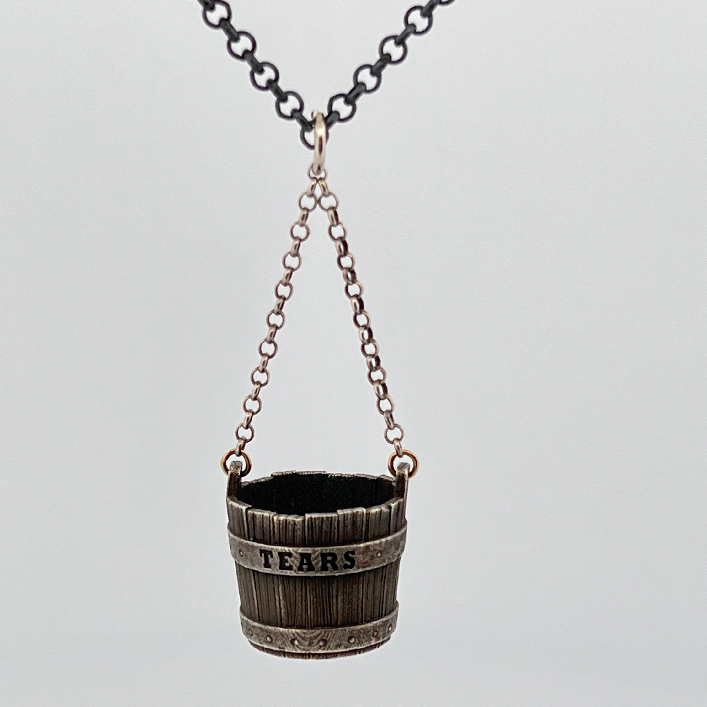 Sterling silver bucket pendant engraved with the word “TEARS,” detailed woodgrain texture, metal band accents, suspended by fine chain links.