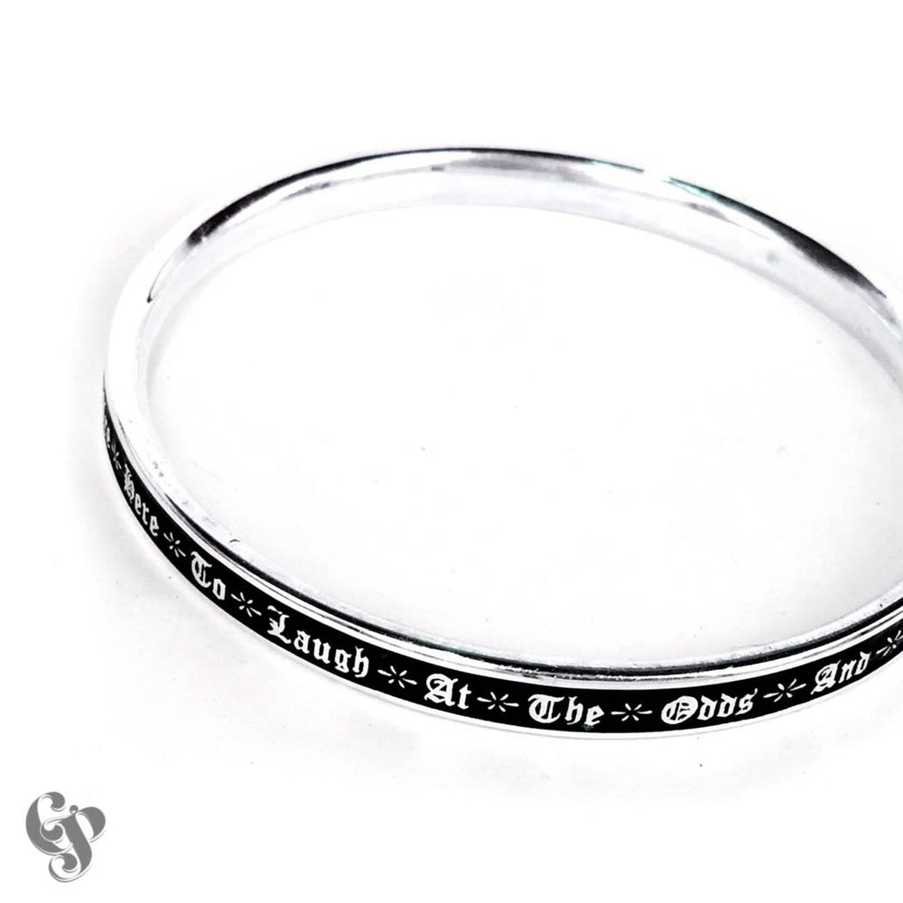 Sterling silver bangle with black enamel engraved gothic script reading “Here to laugh at the odds and…”, slim polished circular band.
