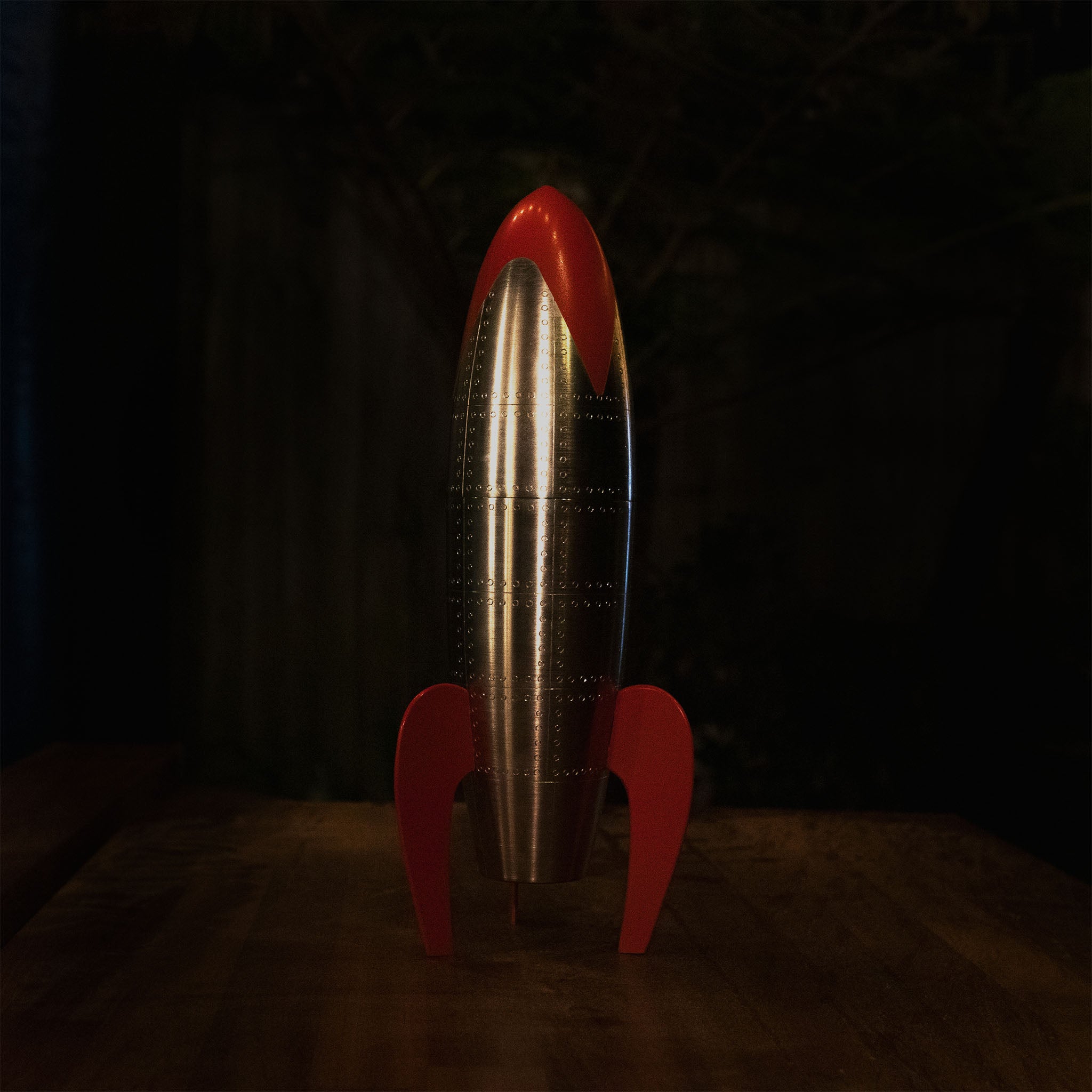 Retro rocket sculpture with polished metal body, riveted panel detailing, and bright red fins and nose cone, standing upright in classic mid-century sci-fi style.