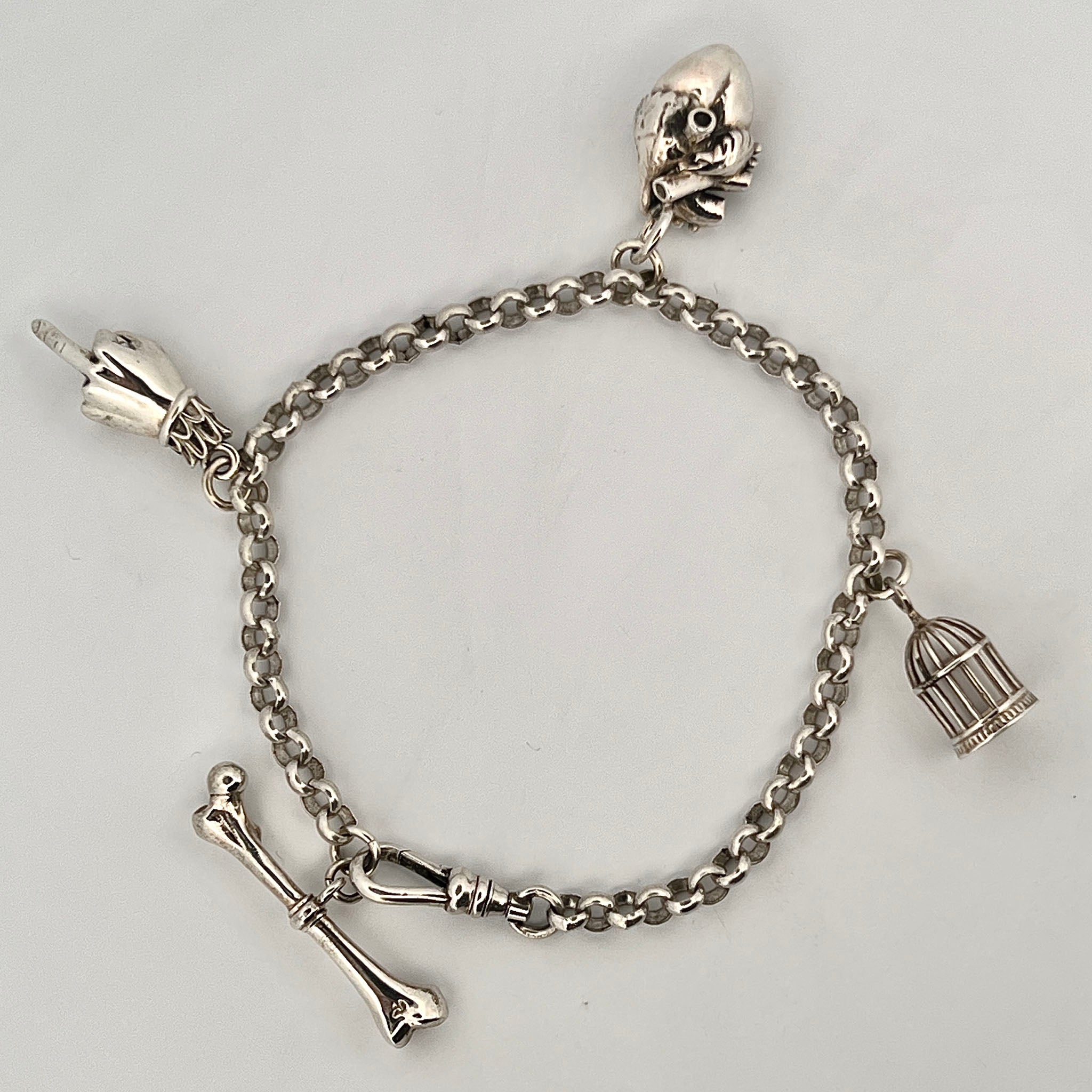 Sterling silver charm bracelet with chunky chain, featuring miniature birdcage charm, detailed bone charm, Victorian hand with raised middle finger charm, and sculpted skull charm.