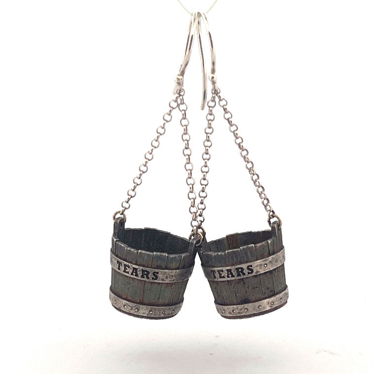 These earrings are crafted in sterling silver and designed as miniature buckets, complete with woodgrain texture and metal banding detail. Each bucket is inscribed with the word TEARS and suspended from fine silver chains on hooks. Measuring approximately 20mm across, they are both sculptural and symbolic, balancing dark humour with fine craftsmanship.