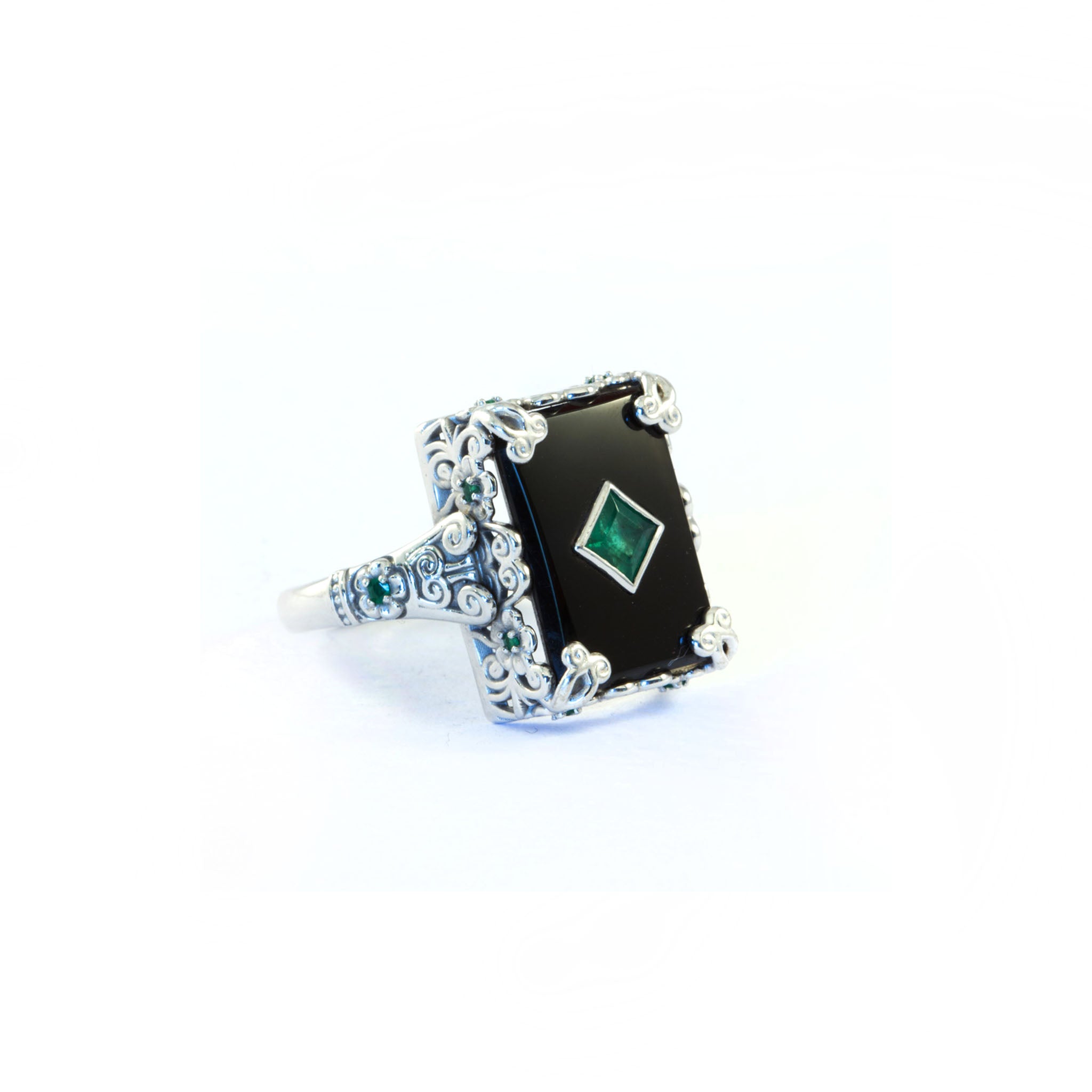 18ct white gold ring with rectangular black onyx panel, central diamond-shaped emerald in bezel setting, ornate scrollwork shoulders set with small round emerald accents.