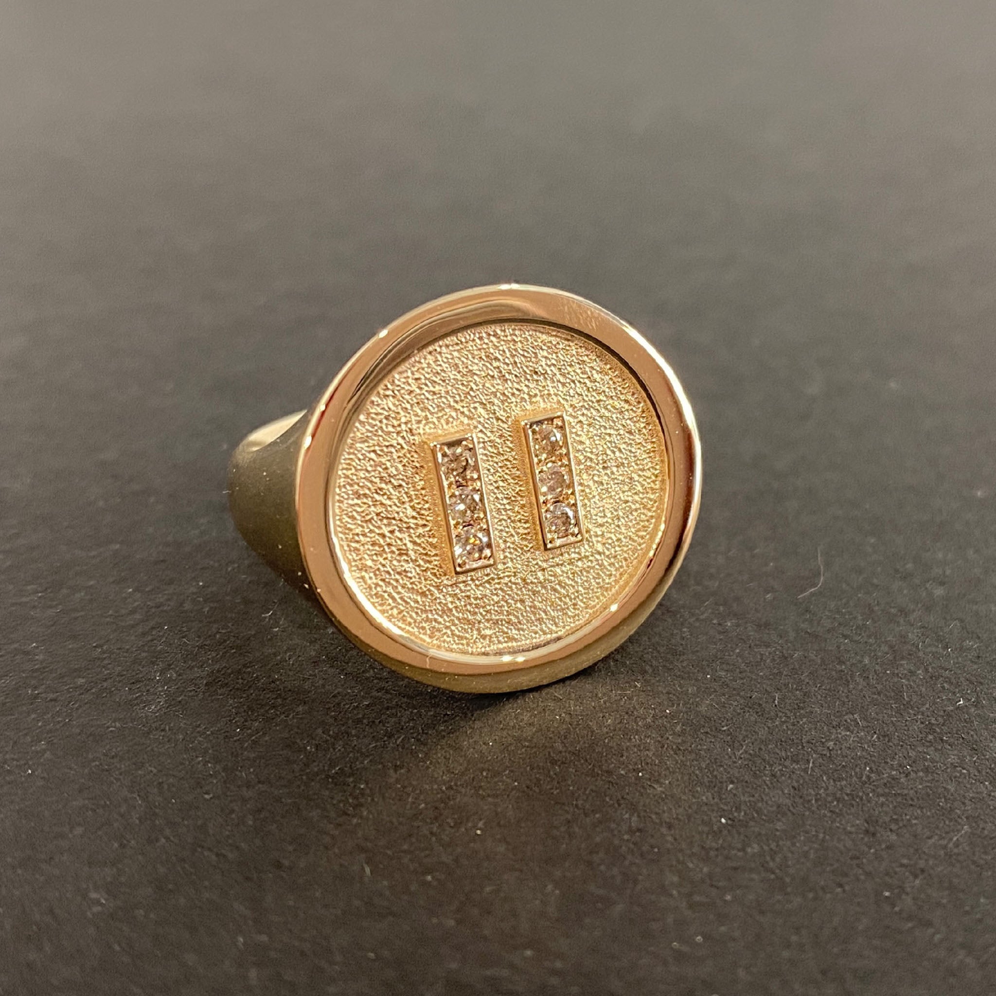 Yellow gold signet-style ring with textured round face and polished rim, featuring two vertical bars pavé-set with round brilliant cut diamonds forming a pause symbol.