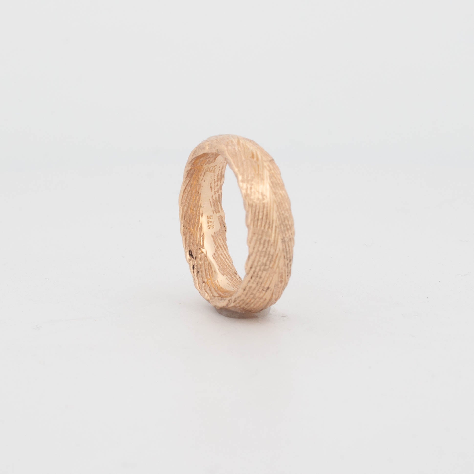 Angled view of a 18ct yellow gold ring cast using a real cuttlefish bone mold, featuring an organic, linear texture with natural grooves and ridges across the band. The surface shows uneven, tactile striations unique to cuttlefish casting, creating a raw, sculptural form with a matte-meets-polished finish.