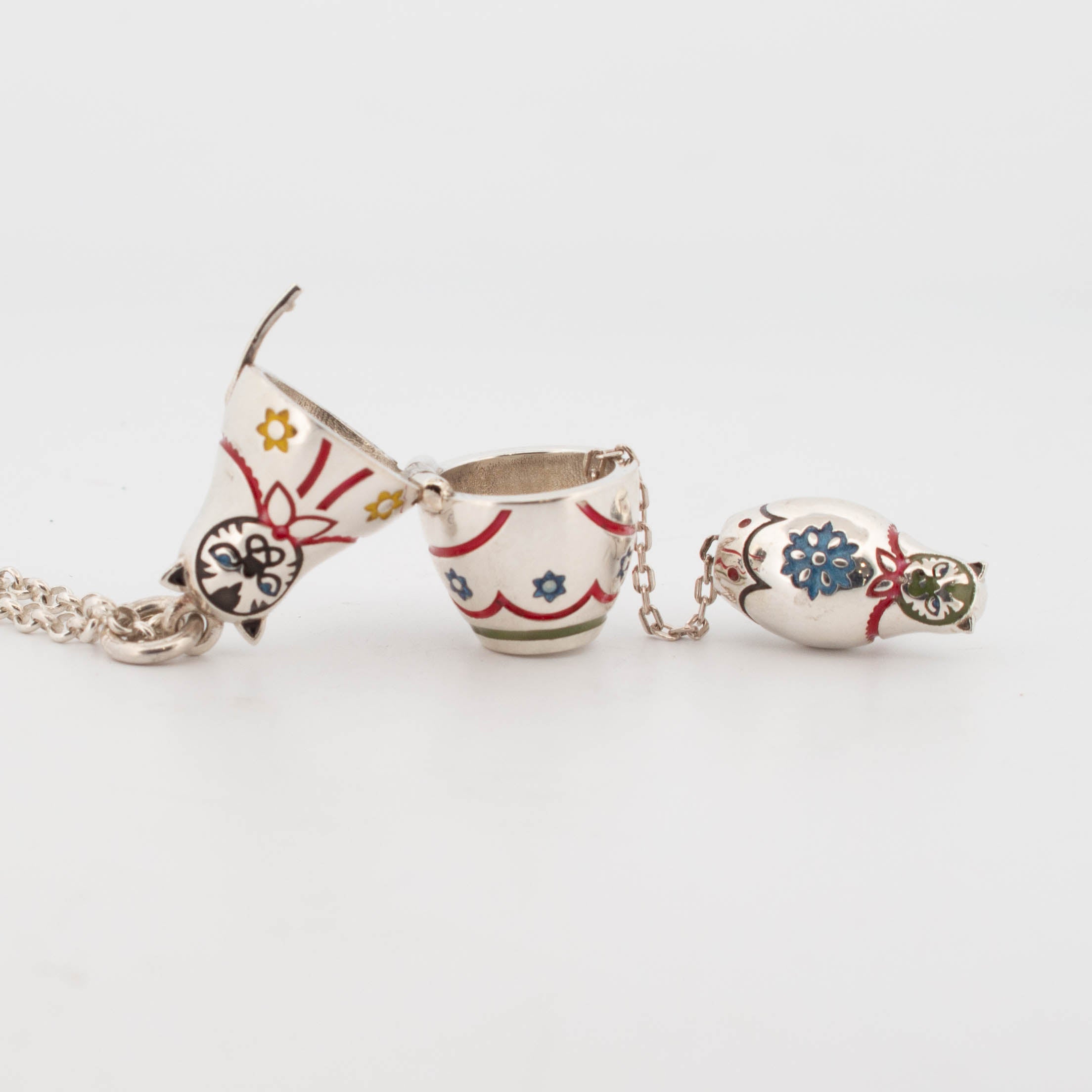Sterling silver hinged babushka pendant shown open to reveal a smaller nested enamel-painted figure inside; both decorated with multicolour floral and ribbon motifs, featuring cat faces and red, yellow, blue, green, and black enamel detailing, attached to a silver chain.