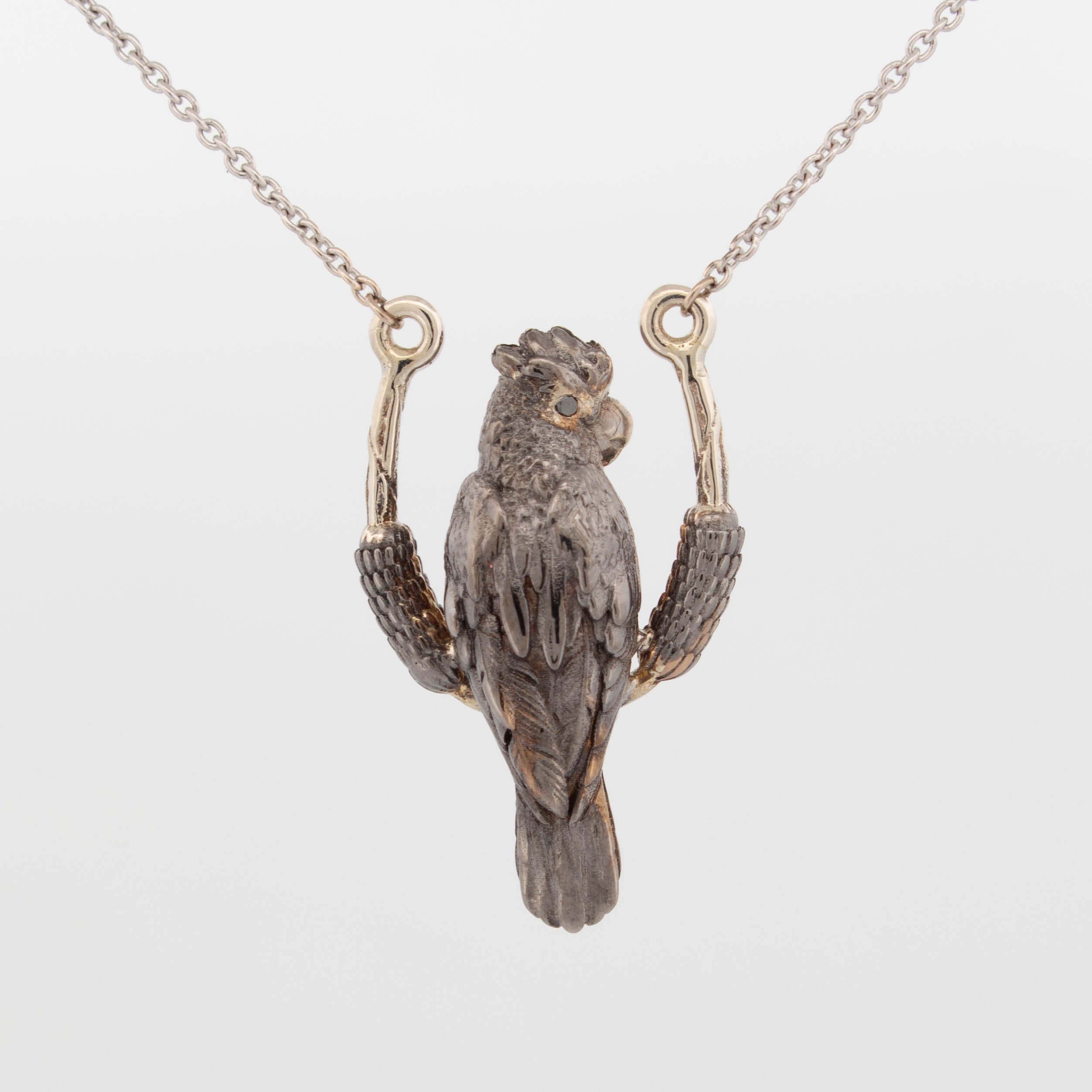 Custom pendant crafted in 9ct white gold with black rhodium plating, depicting a highly detailed black cockatoo perched on a curved branch, with black diamond eyes and a polished 9ct yellow gold cheek highlight; suspended on a fine gold chain.