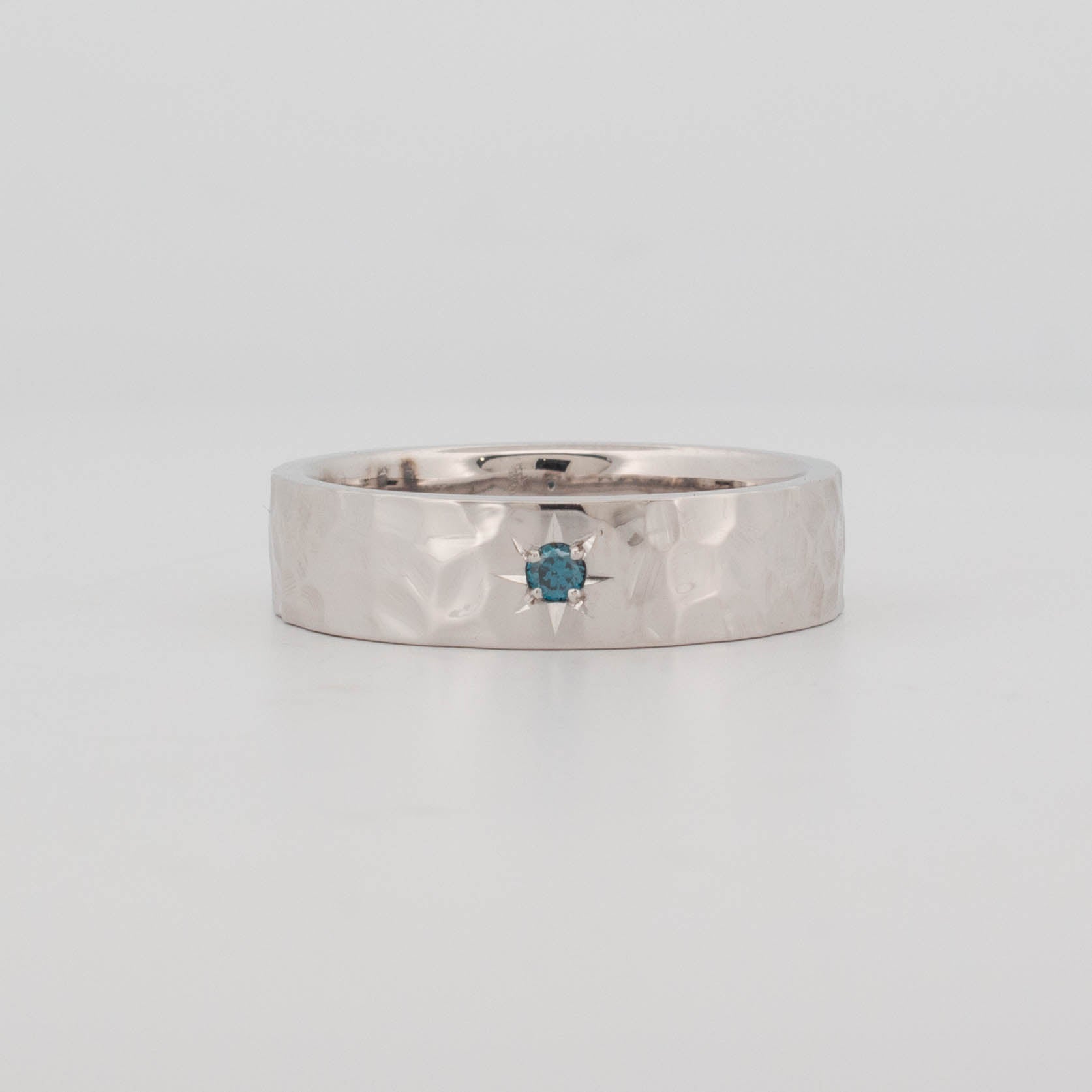 Front view of a cobblestone-textured 9ct white gold ring, featuring a centrally set irradiated blue diamond in a hand-engraved star setting. The band has a high-polish, hammered surface resembling natural stone, offering a tactile and organic finish. A modern ring design with artisanal detail and a vibrant blue focal stone.