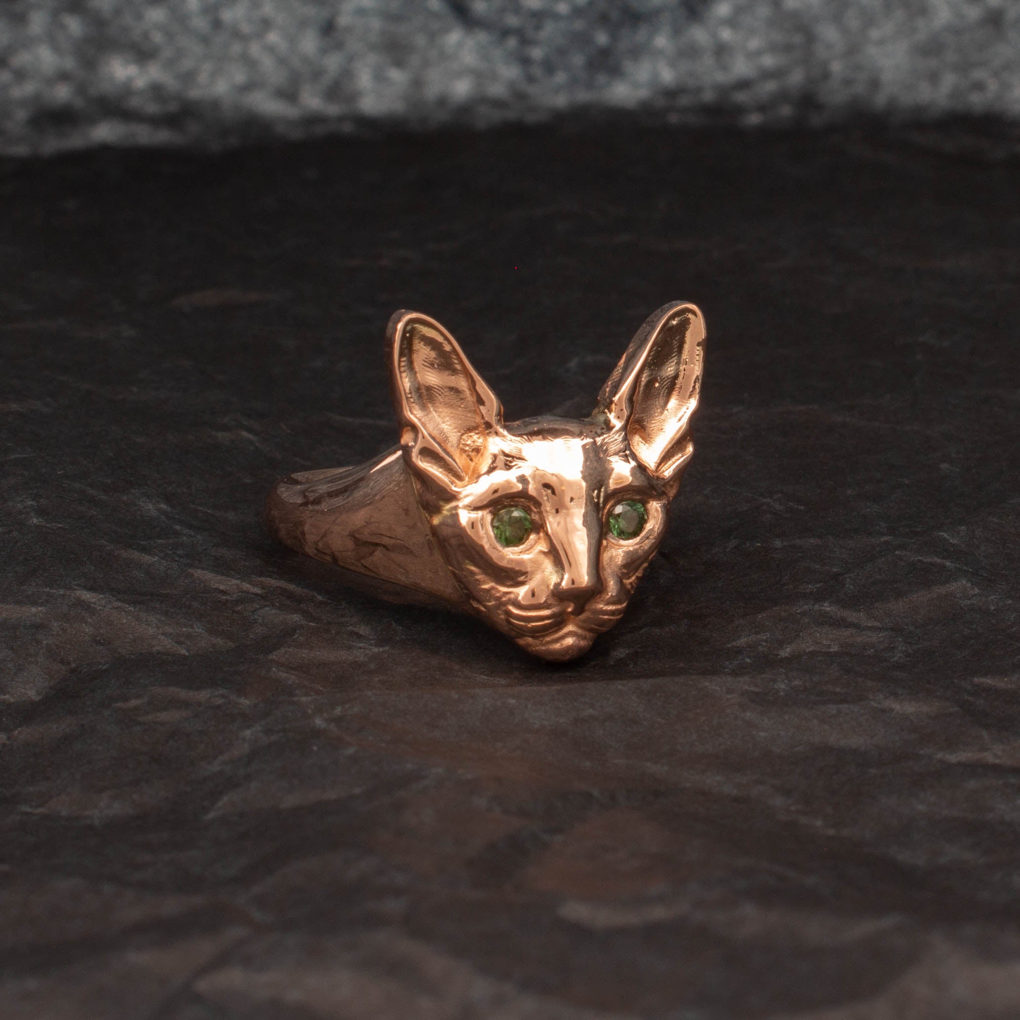9ct yellow gold ring featuring a sculpted Sphynx cat head with detailed ears and whiskers, set with round green sapphires as eyes; customisable with alternative gemstone colours.
