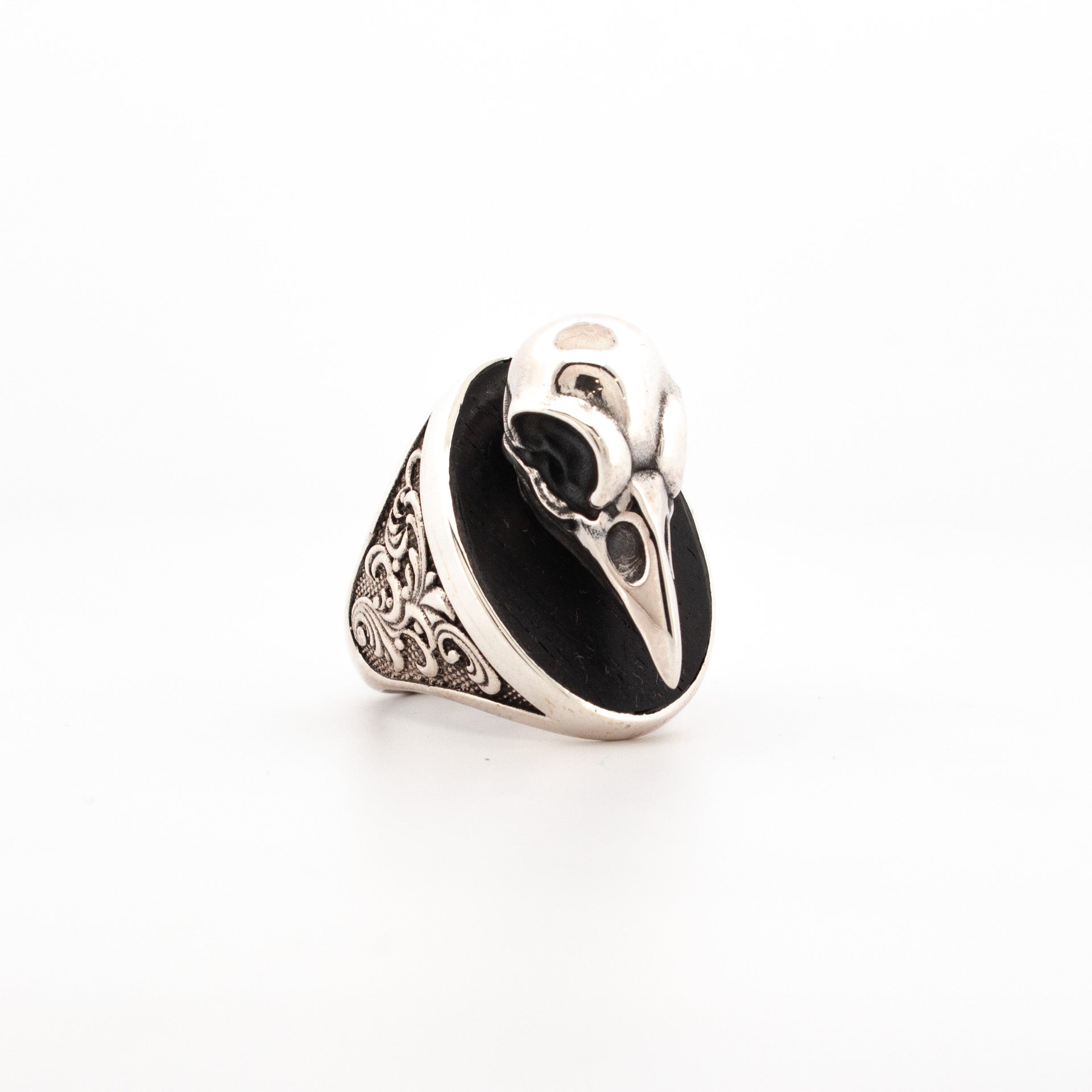 Sterling silver ring featuring sculpted raven skull over oval ebony inlay with engraved scrollwork shoulders.