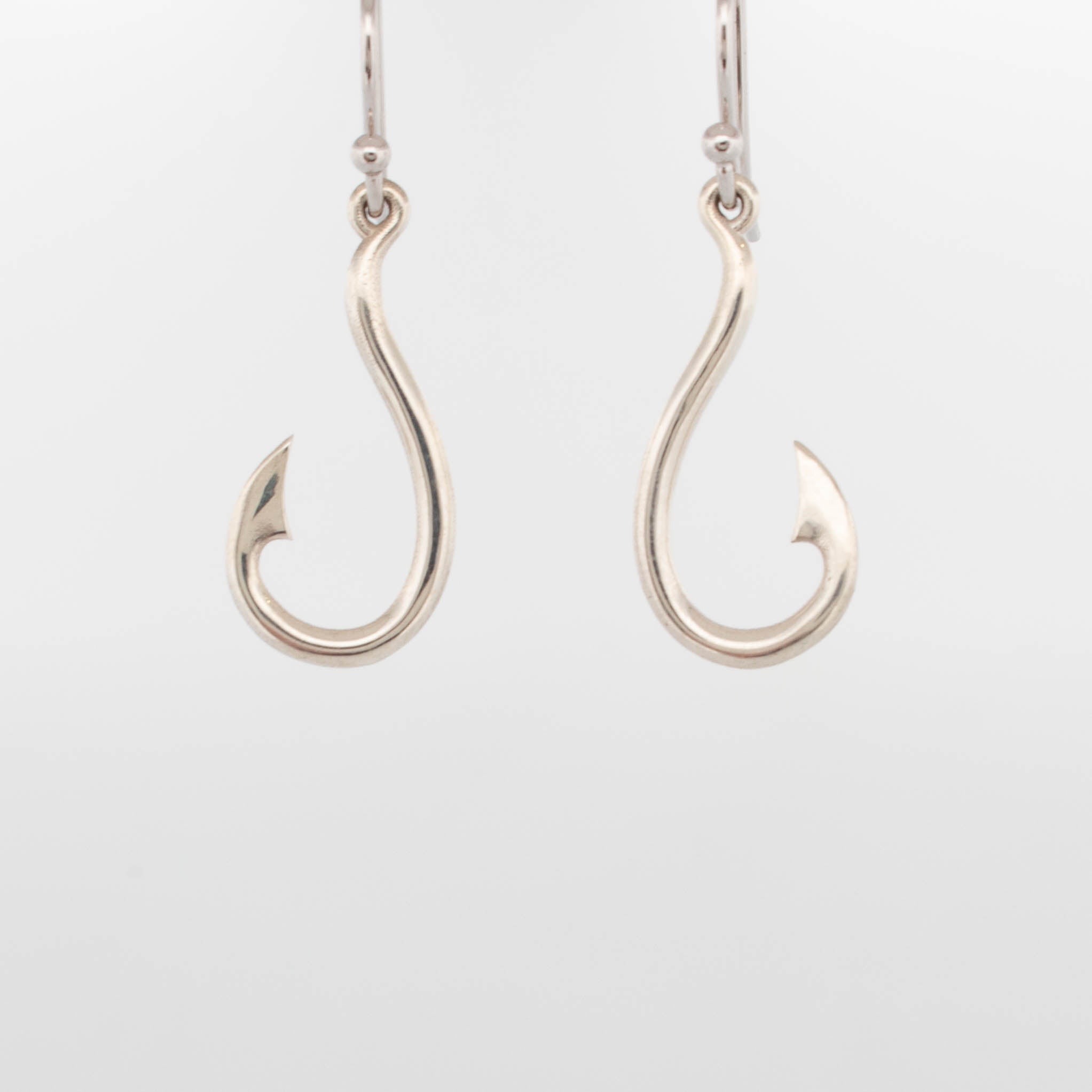 Pair of sterling silver fish hook earrings with a polished finish, shaped like curved fishing hooks with pointed barbs, suspended from classic ear wires; minimalist, nautical-inspired jewellery design photographed on a white background.