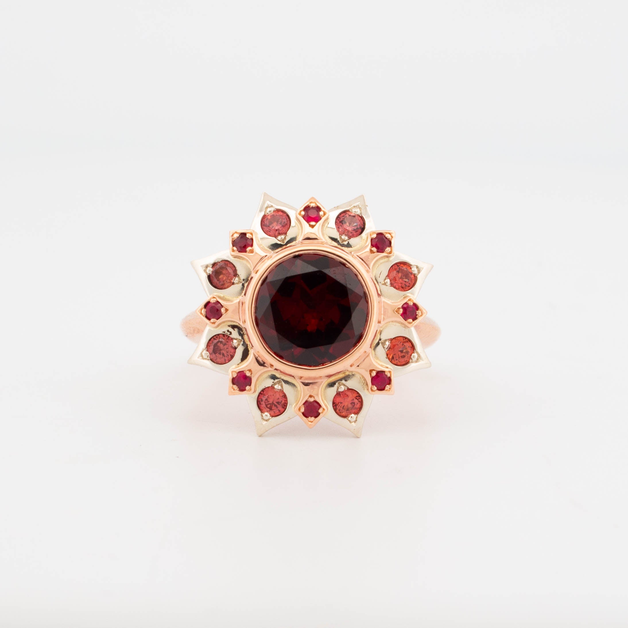 Front view of 9ct rose gold ring featuring a large round garnet at the centre, surrounded by flame-shaped settings with red rubies and pink sapphires in a sunburst arrangement; handcrafted gemstone cluster design with warm tonal palette.