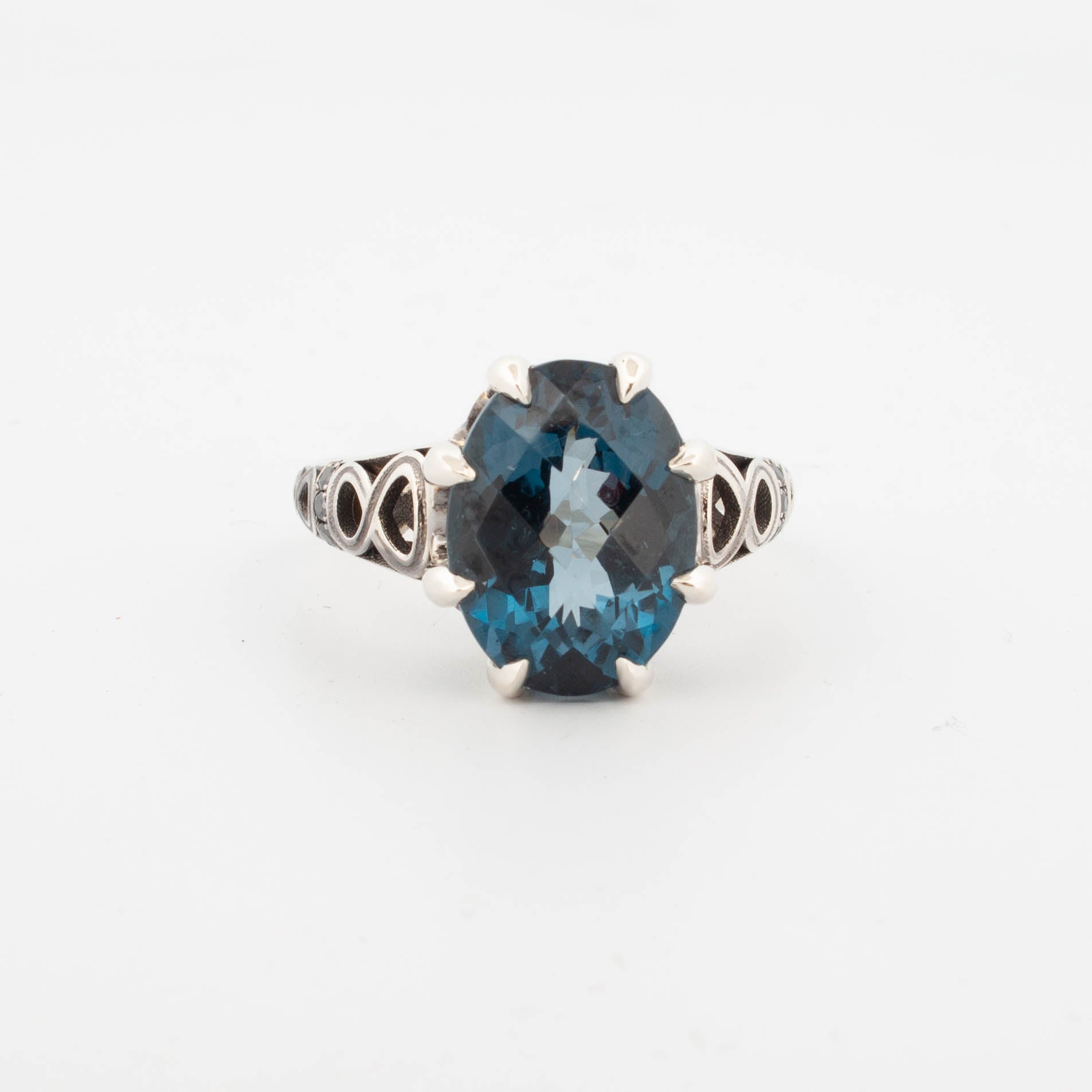 9ct white gold ring featuring a large oval-cut London Blue Topaz in a claw setting, with infinity symbol openwork shoulders and pavé-set black diamonds on the band.