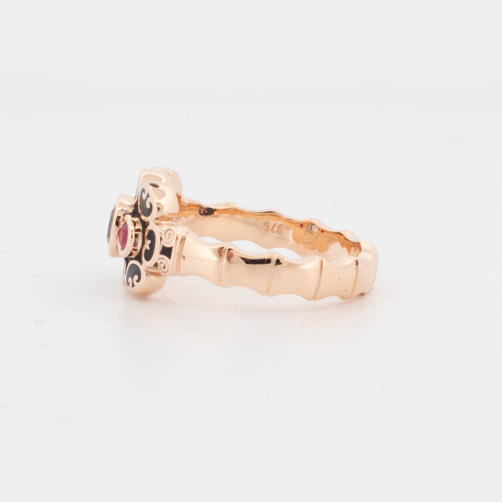 9ct rose gold ring with black enamel scrollwork, featuring a central cushion-cut blue-green sapphire bezel-set, flanked by two round-cut rubies in polished settings.