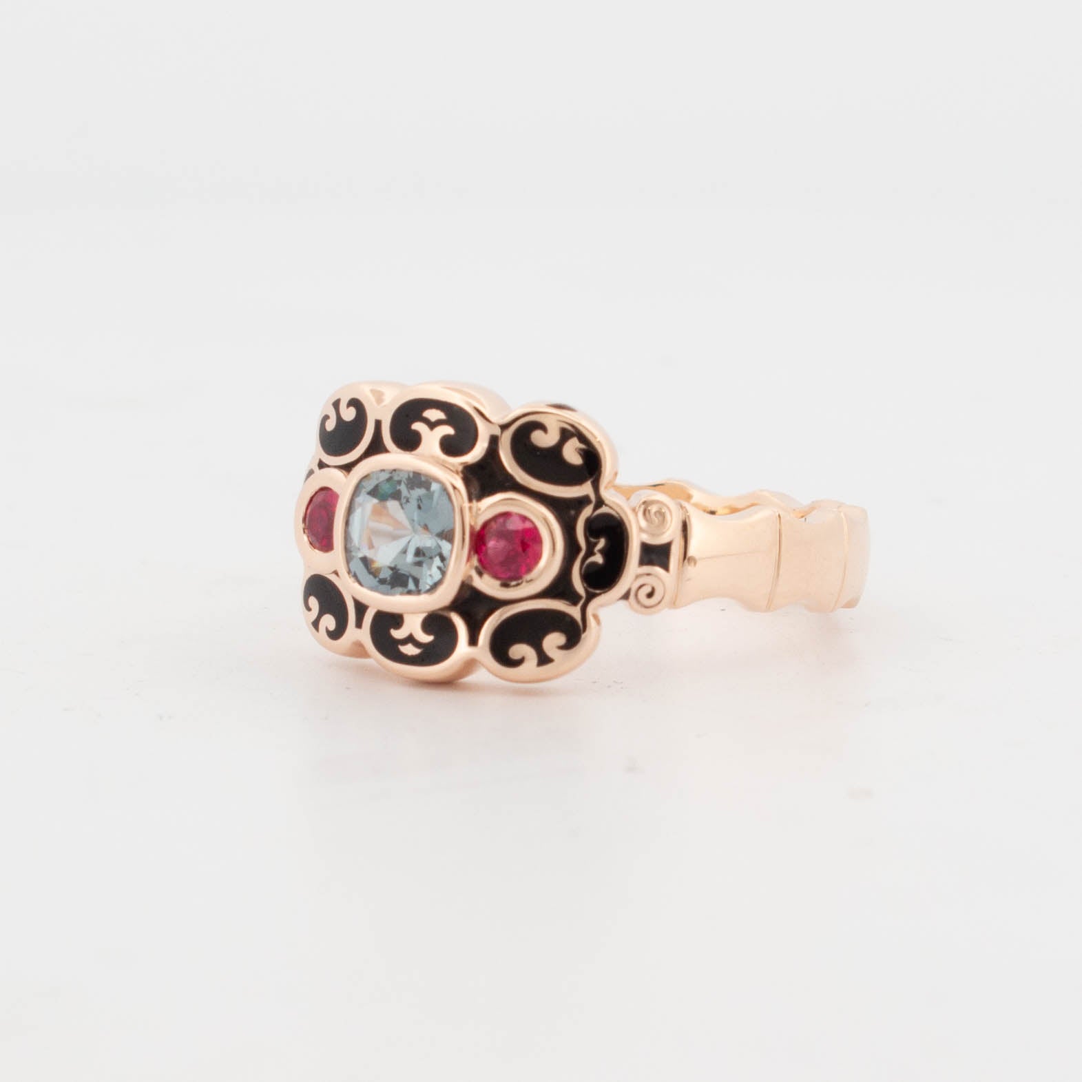 9ct rose gold ring with black enamel scrollwork, featuring a central cushion-cut blue-green sapphire bezel-set, flanked by two round-cut rubies in polished settings.