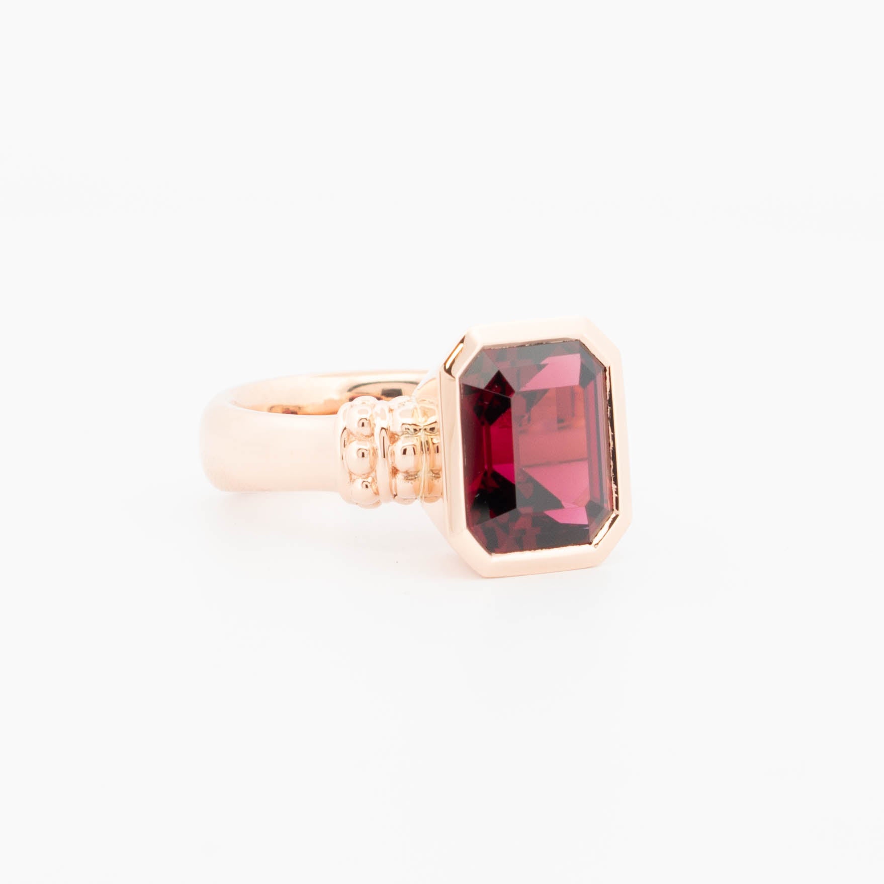 18ct rose gold ring featuring a richly coloured emerald-cut Rhodolite Garnet in a bezel-style setting, with a bold, polished band and decorative beaded shoulder detail.