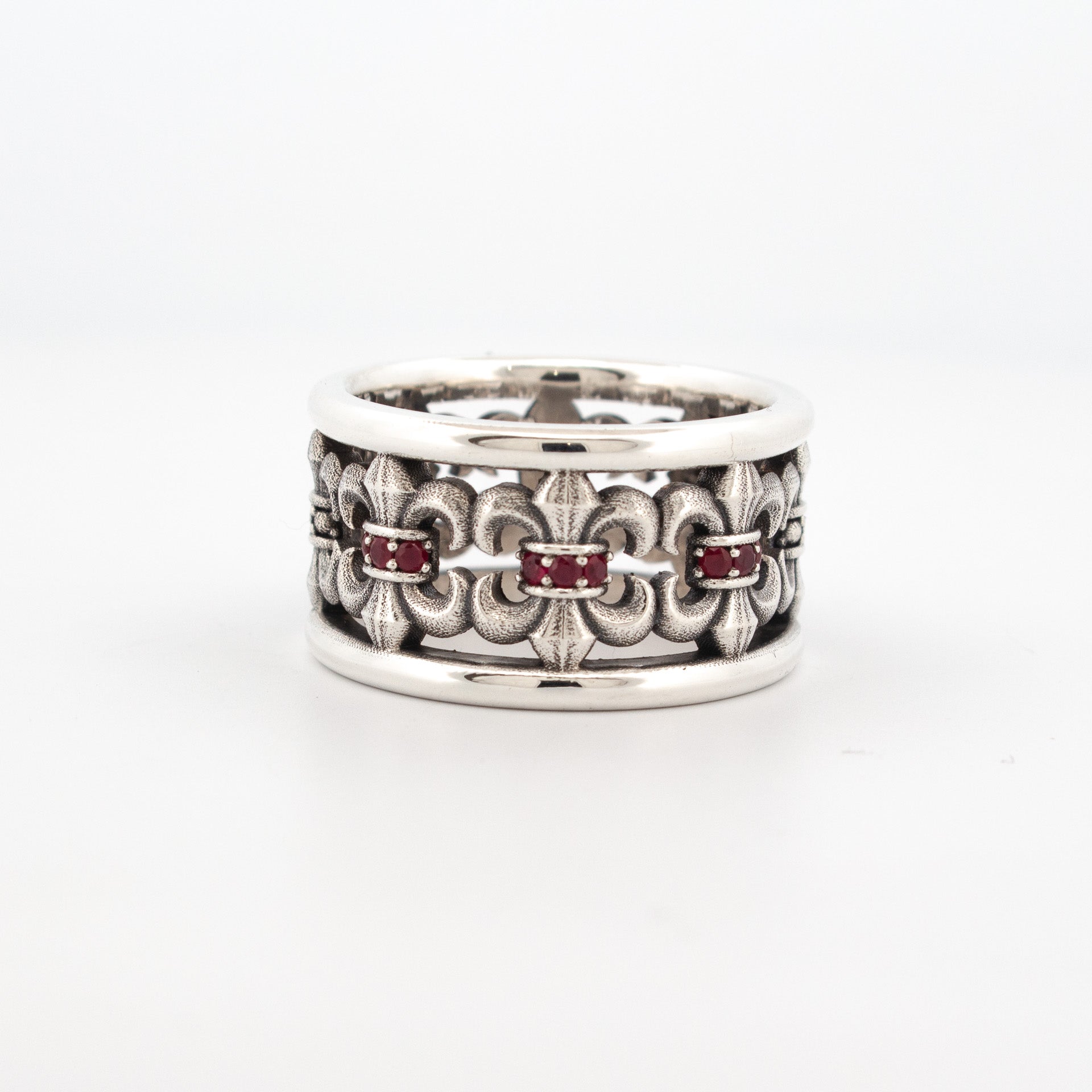 Sterling silver wide band ring with repeating fleur-de-lis motif and bezel-set red rubies.