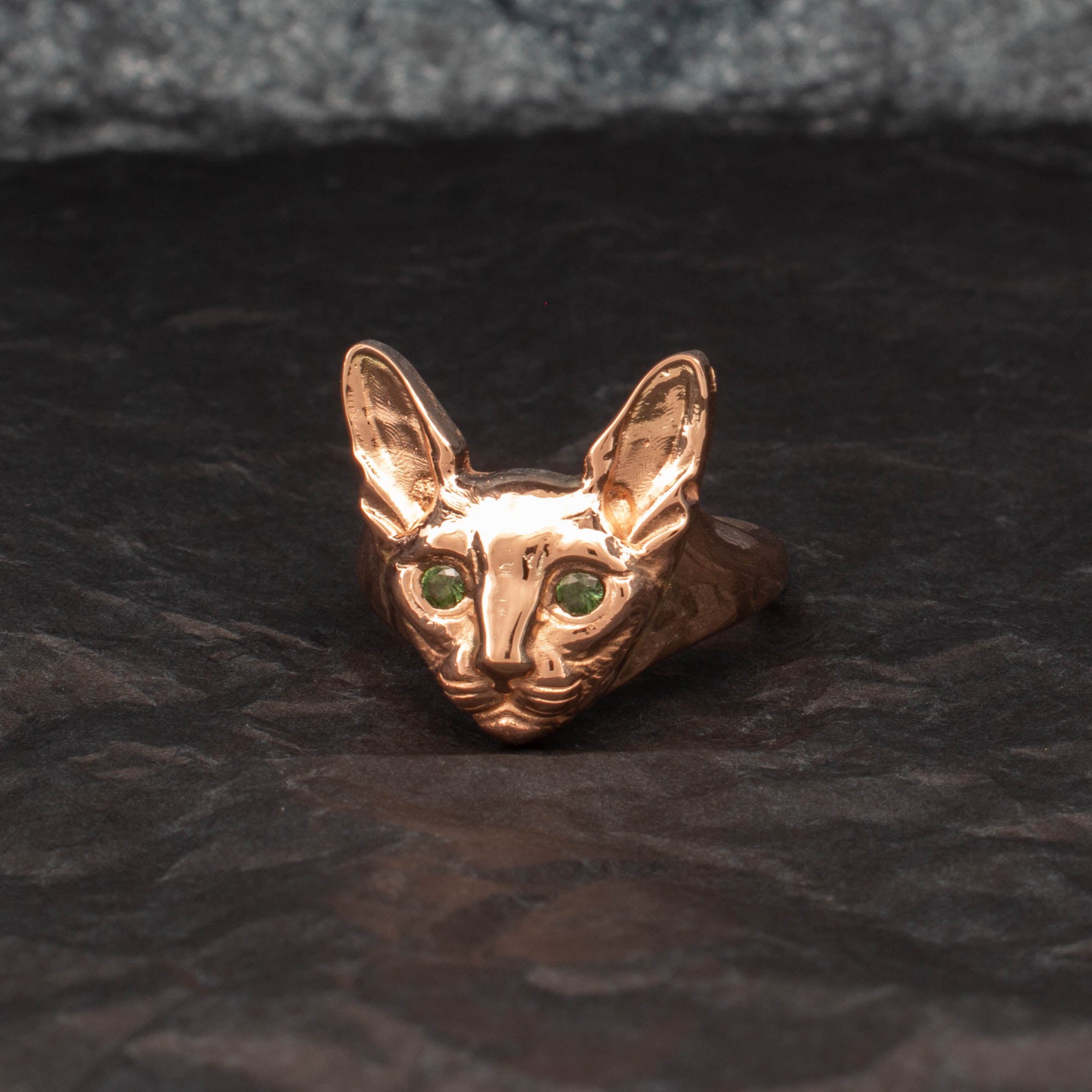 9ct yellow gold ring featuring a sculpted Sphynx cat head with detailed ears and whiskers, set with round green sapphires as eyes; customisable with alternative gemstone colours.