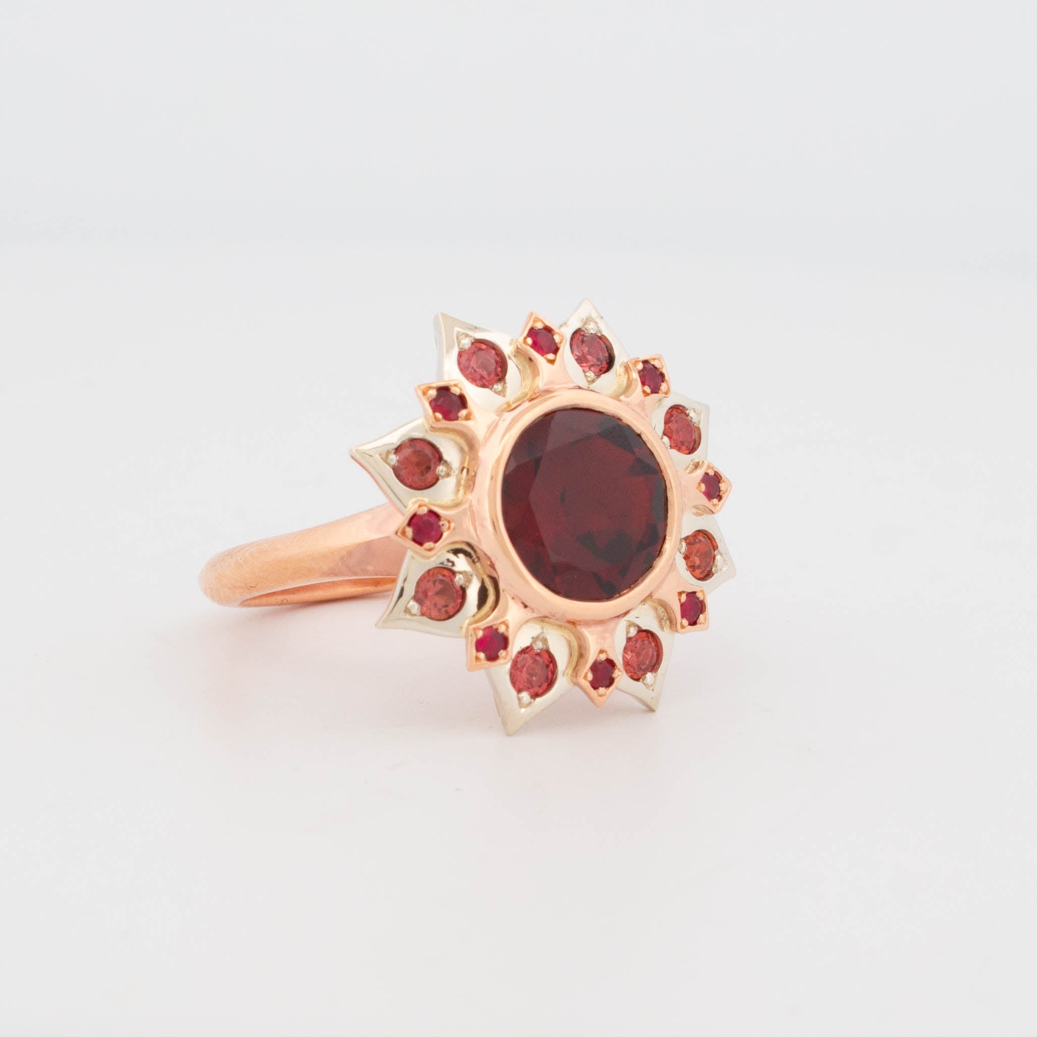 Side view of 9ct rose gold ring featuring a large round garnet at the centre, surrounded by flame-shaped settings with red rubies and pink sapphires in a sunburst arrangement; handcrafted gemstone cluster design with warm tonal palette.