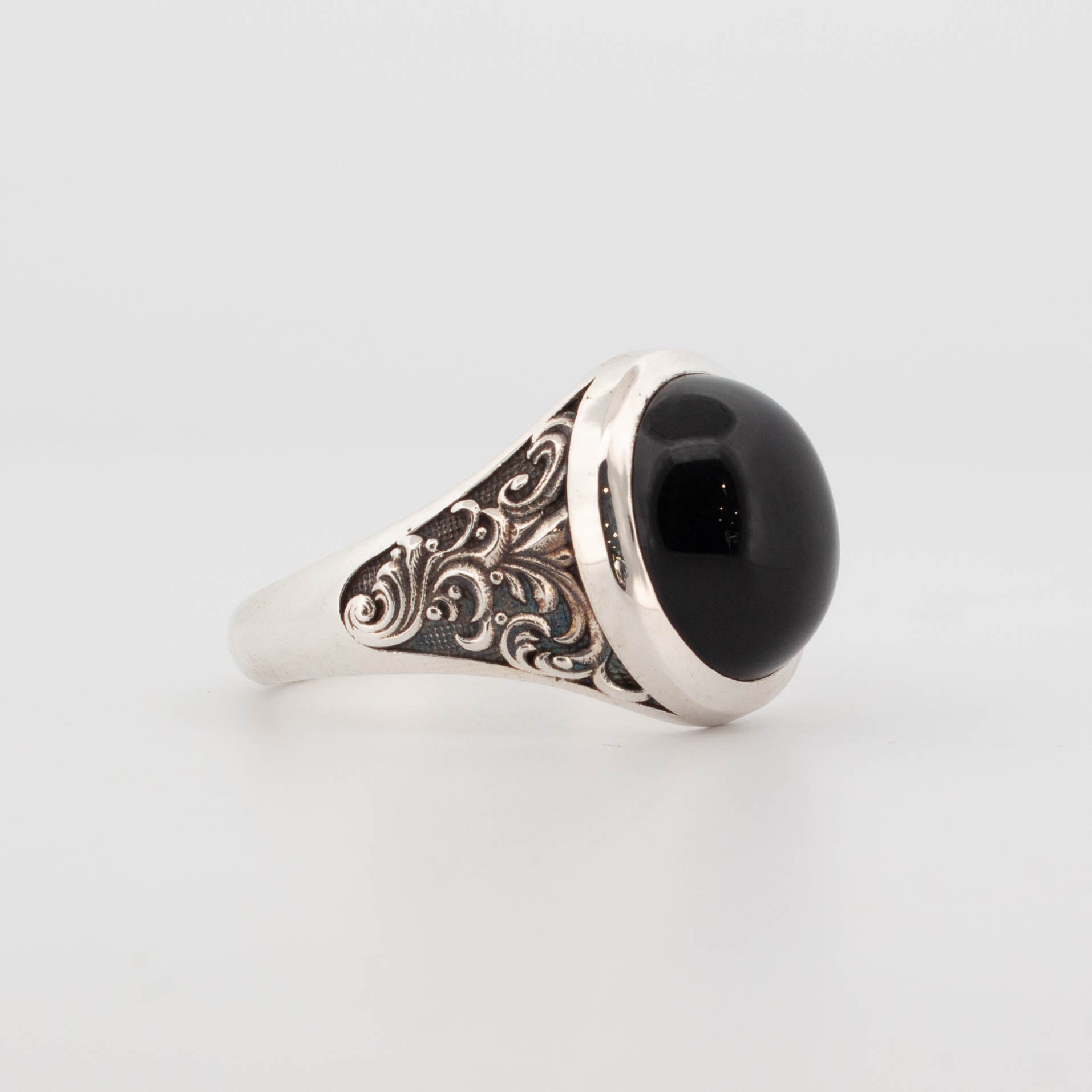 Side view of a large sterling silver ring featuring an oval cabochon black onyx set in a smooth bezel. The tapered band showcases ornate, oxidised scrollwork with intricate filigree detailing, creating a rich vintage or gothic look. The domed onyx adds a bold, polished finish, making it a standout piece for fans of statement rings and handcrafted gemstone jewellery.