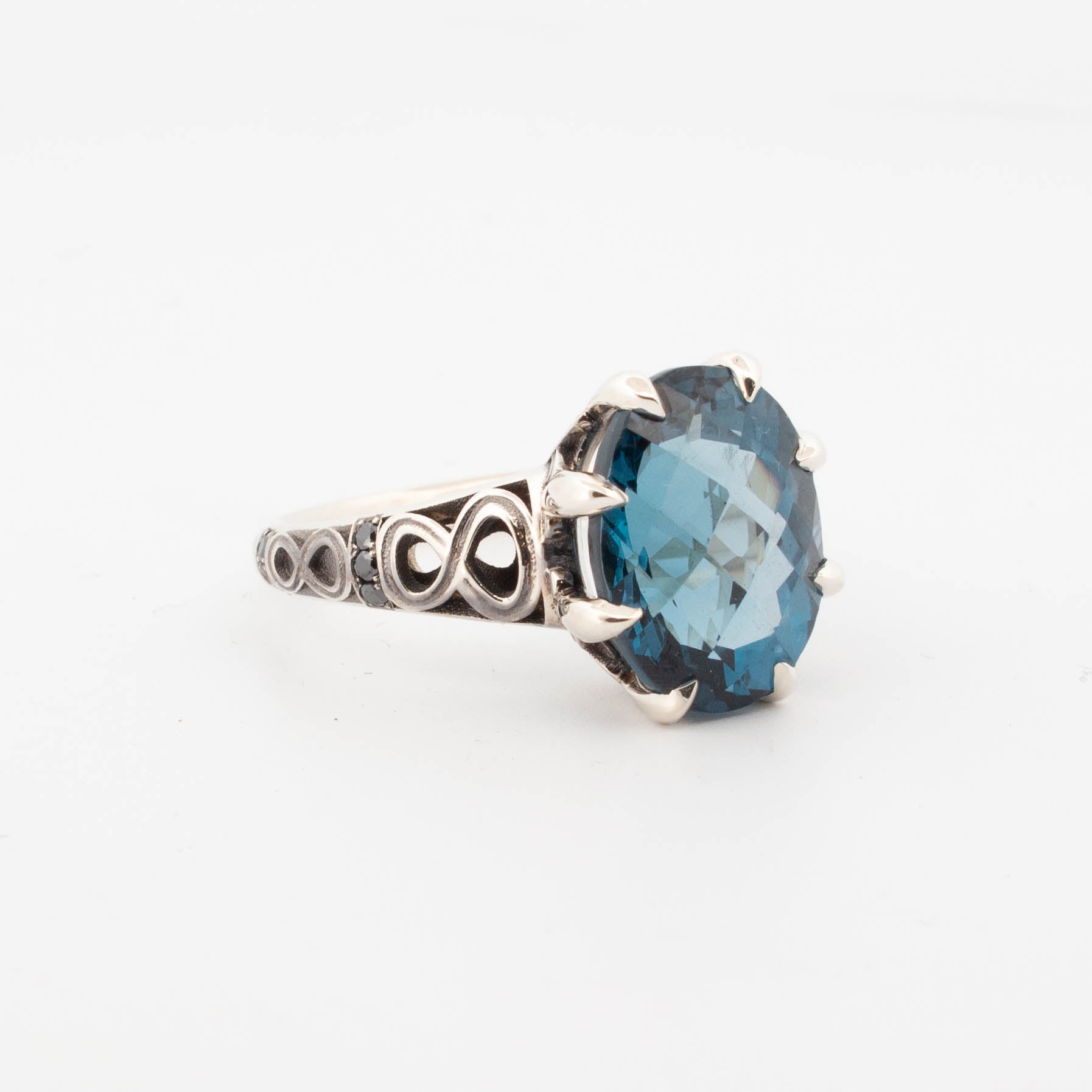 9ct white gold ring featuring a large oval-cut London Blue Topaz in a claw setting, with infinity symbol openwork shoulders and pavé-set black diamonds on the band.