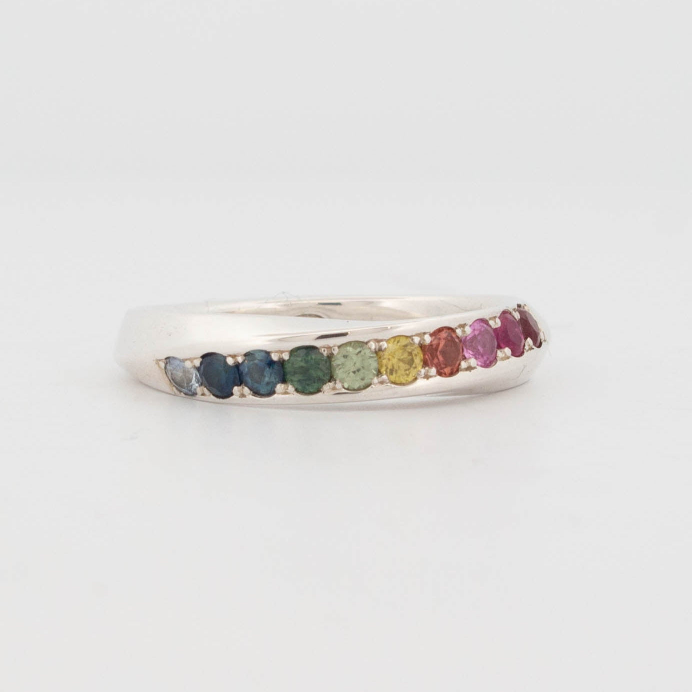 Sterling silver rainbow sapphire ring featuring a smooth half-round band set with a gradient of round-cut sapphires in a seamless colour fade—from deep blue and teal to green, yellow, orange, pink, and red. The flush pavé-style setting enhances the sleek, modern look while showcasing the full spectrum of natural sapphire hues. A minimalist yet vibrant design perfect for pride jewellery, colourful stacking rings, or everyday gemstone bands.