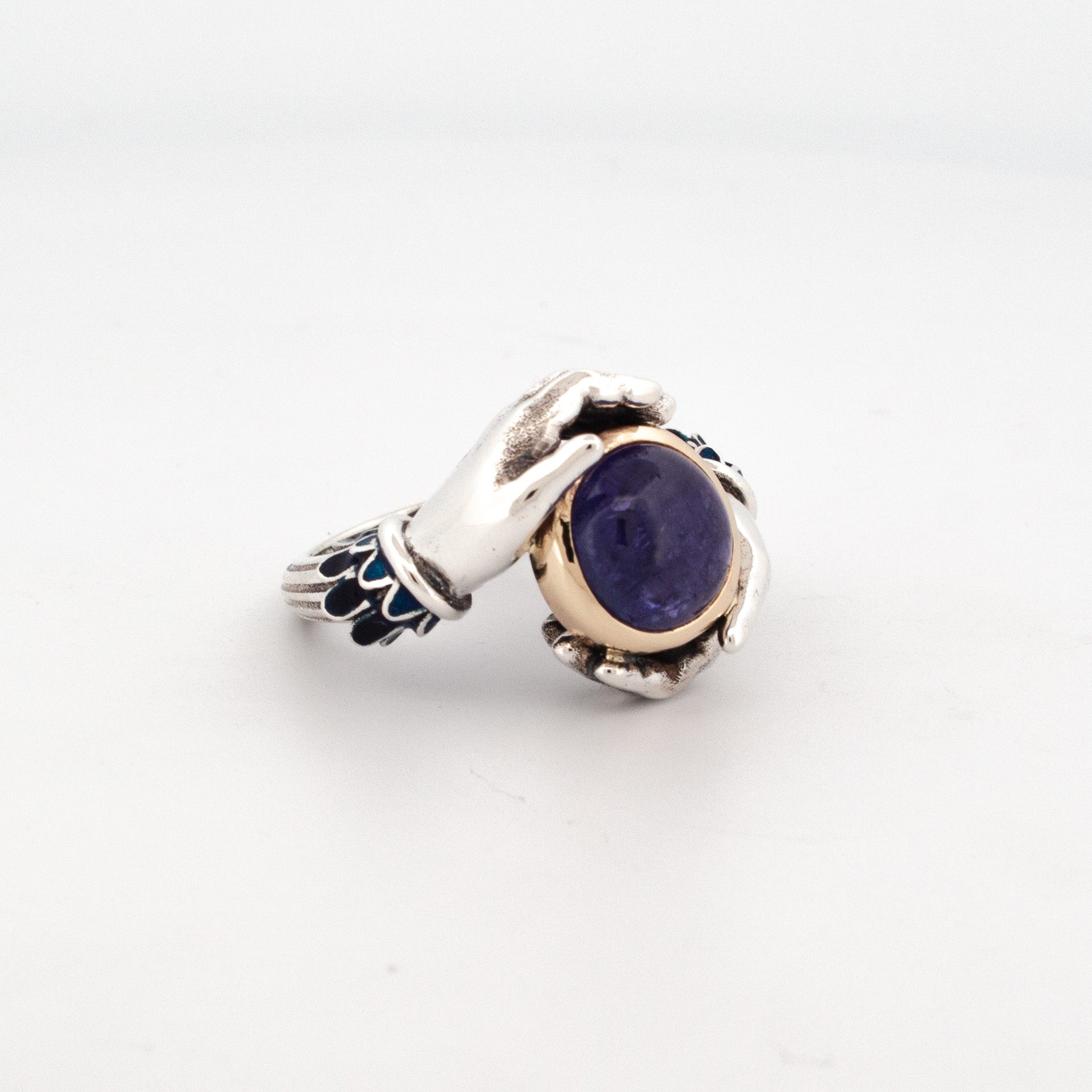 side view Sterling silver ring with enamelled sleeves and 9ct yellow gold bezel set 10mm round cabochon tanzanite, held by sculpted hands.