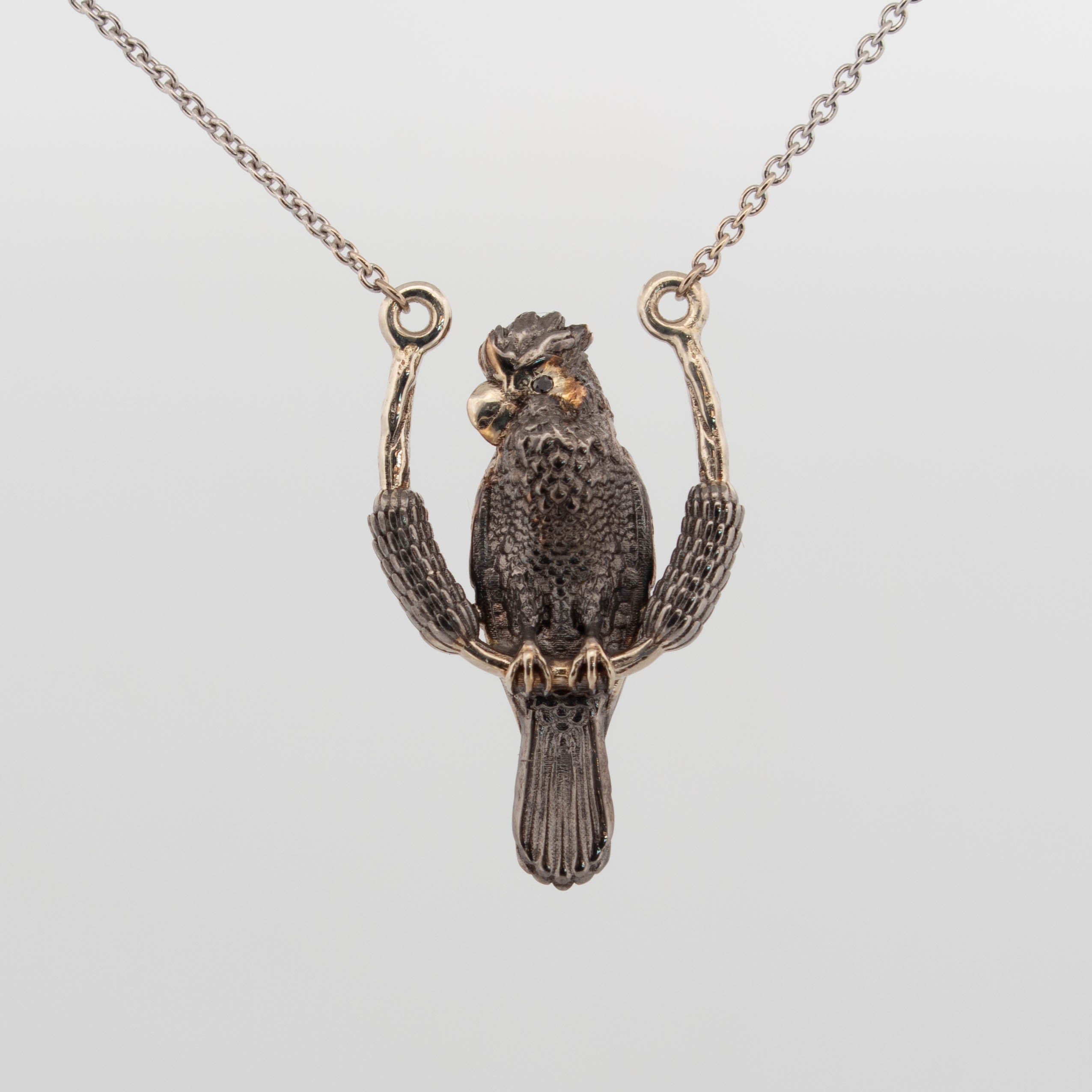 Custom pendant crafted in 9ct white gold with black rhodium plating, depicting a highly detailed black cockatoo perched on a curved branch, with black diamond eyes and a polished 9ct yellow gold cheek highlight; suspended on a fine gold chain.