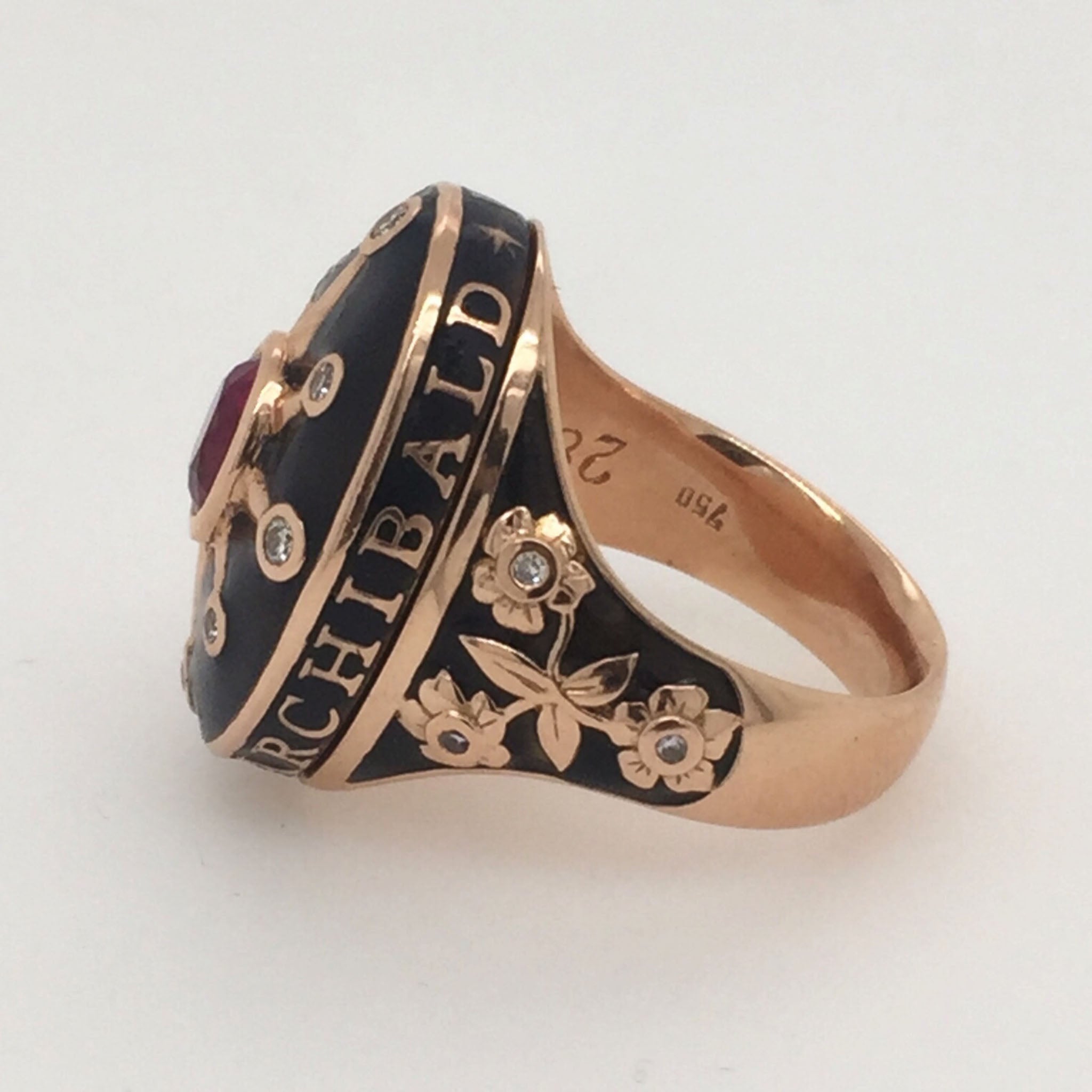 A commemorative ring with a black enamel forget-me-not flower detail, featuring a ruby centerpiece and encrusted with diamonds. The ring has a rose gold band with the name 'Archibald' inscribed on it.