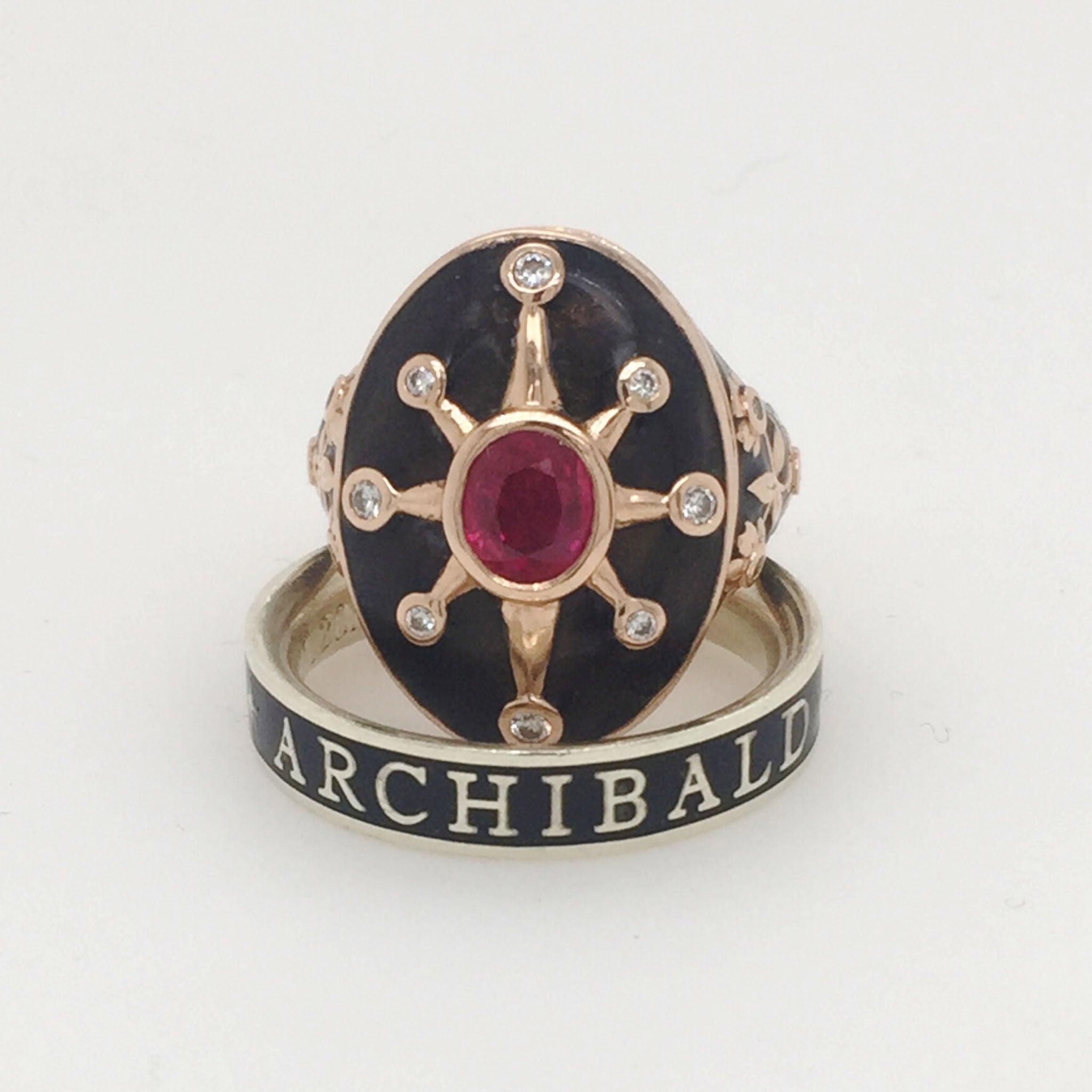 A commemorative ring with a black enamel forget-me-not flower detail, featuring a ruby centerpiece and encrusted with diamonds. The ring has a rose gold band with the name 'Archibald' inscribed on it.