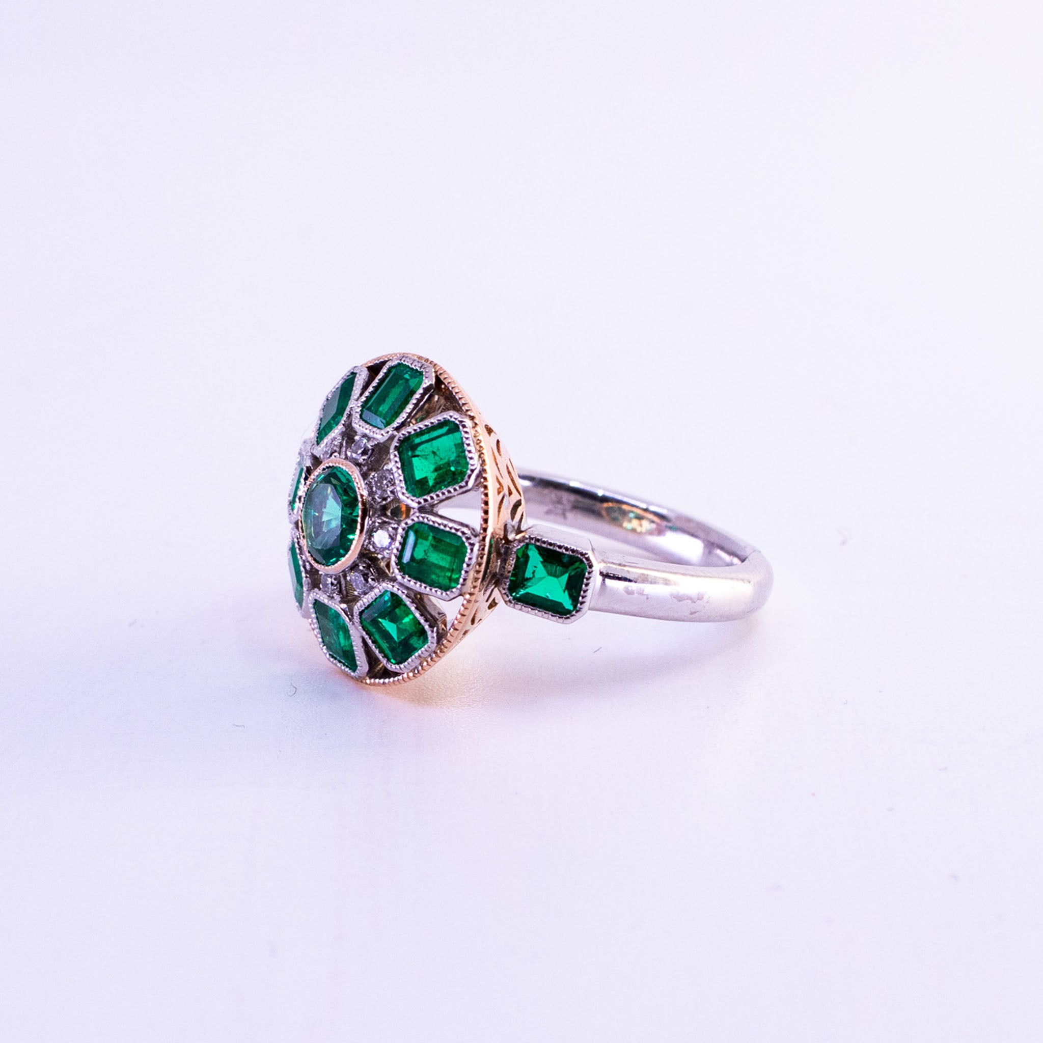 Custom 18ct rose and white gold Art Deco ring featuring central round emerald and halo of square-cut Colombian emeralds with round diamond accents and detailed geometric setting.